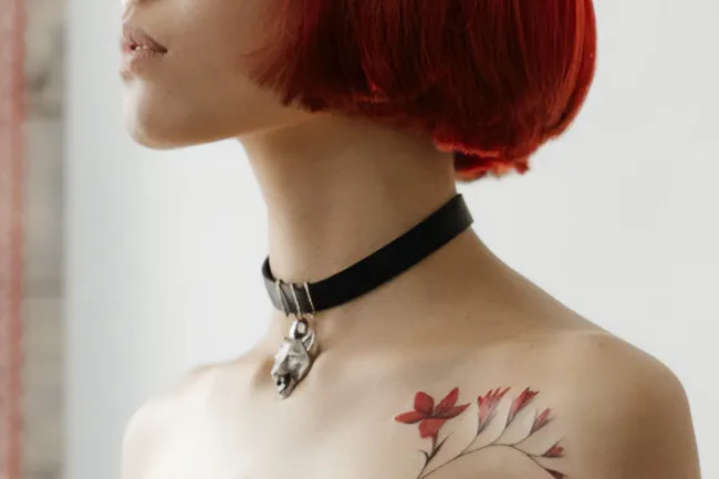 A woman with red hair wearing a choker with a metallic wolf head charm. She has a red floral tattoo.