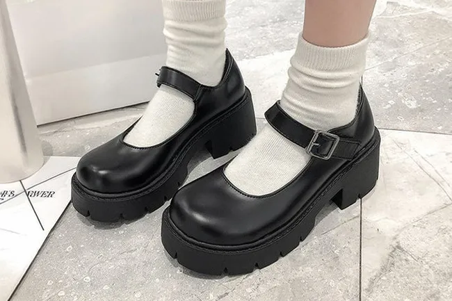 A person wearing black platform Mary Jane shoes with buckles and white scrunched socks on a light gray floor.