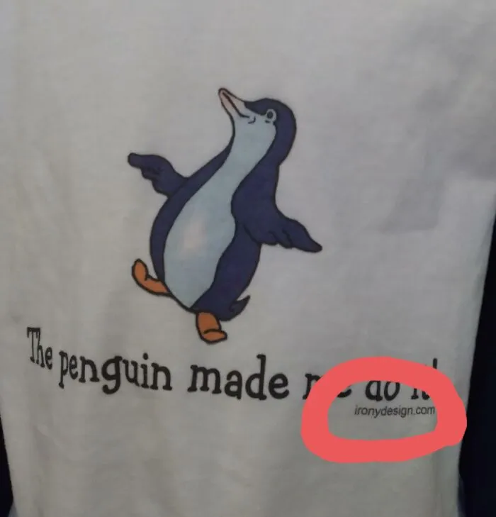 A white t-shirt with a penguin illustration and bad clothing designs text that says 'The penguin made r' with 'ironydesign.com' circled.