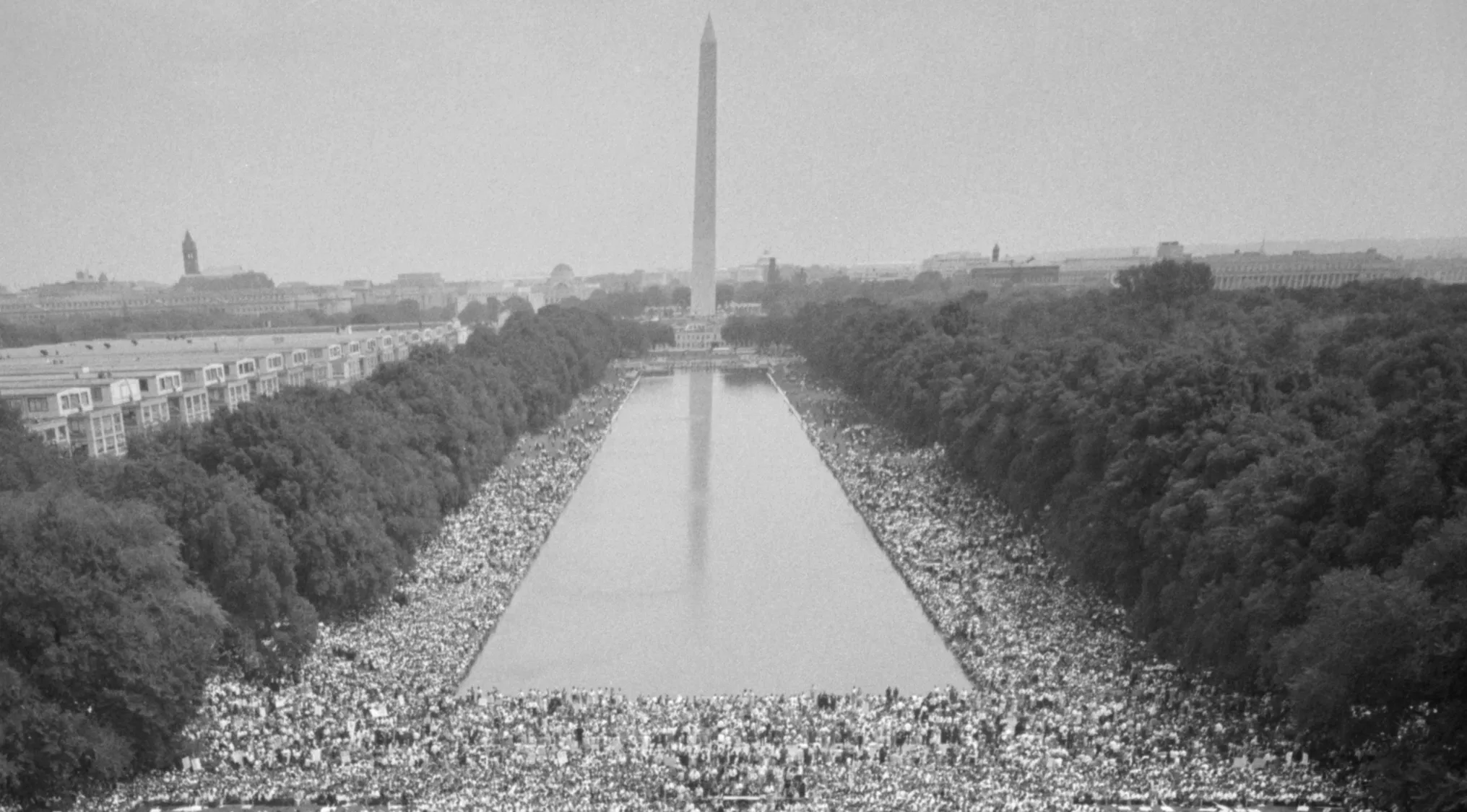 Black and white aerial view of the March on Washington, with crowds lining the reflecting pool, focusing on the I Have a Dream speech.