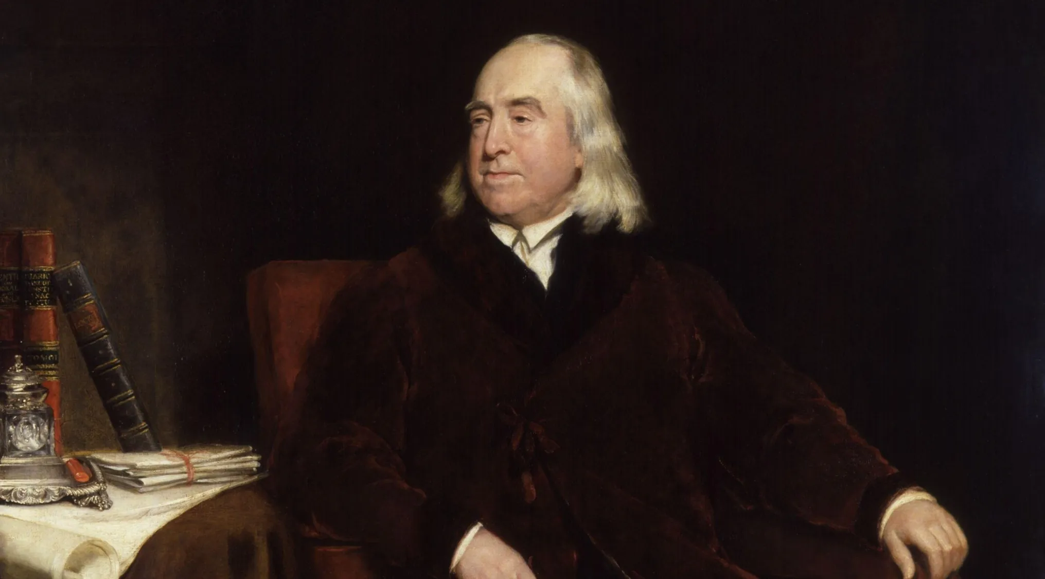 Portrait of Jeremy Bentham, a key figure in utilitarianism, seated in a red robe with books beside him.