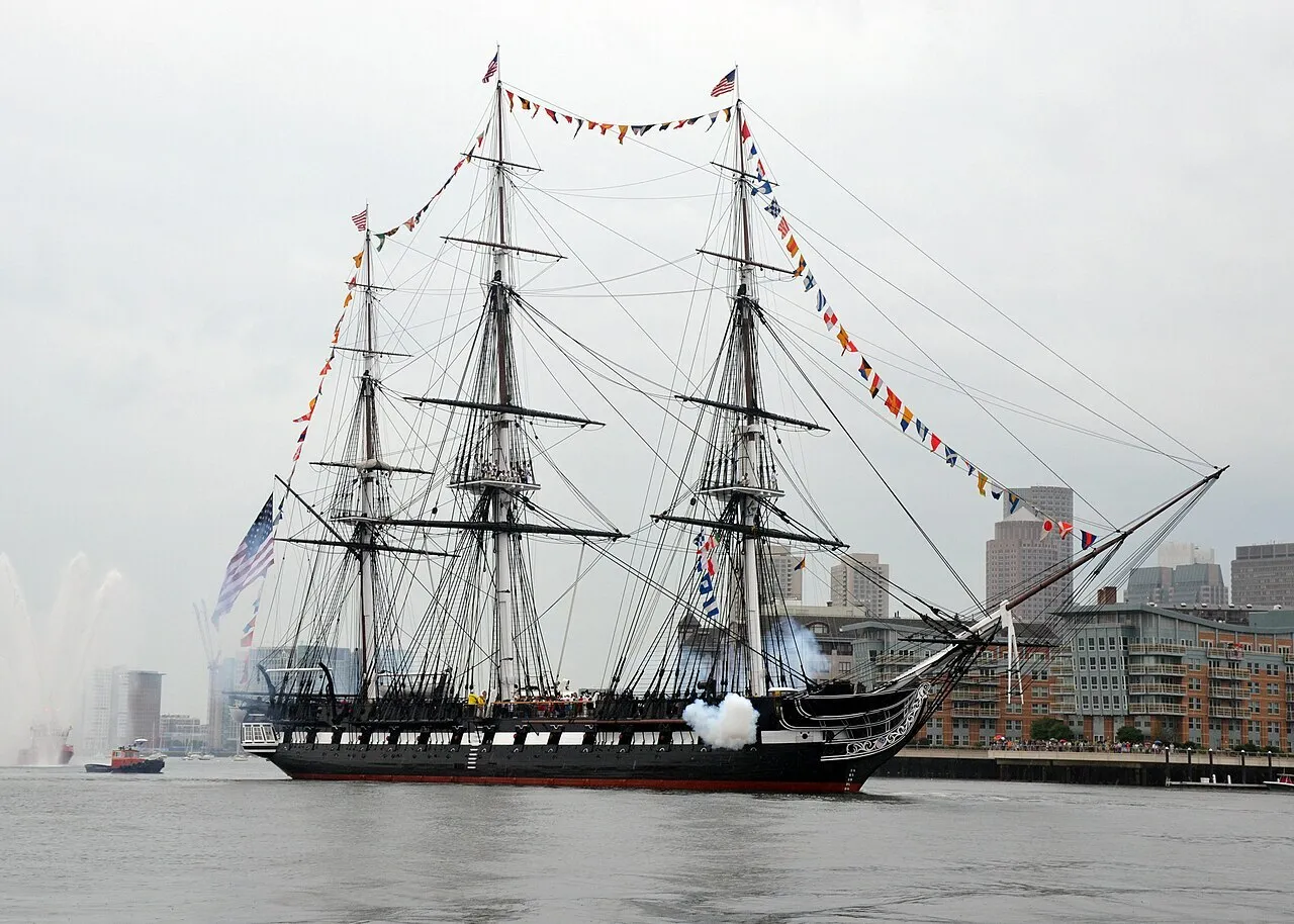 The Ship of Theseus, a historic frigate firing a cannon, sails past a city skyline on a cloudy day.