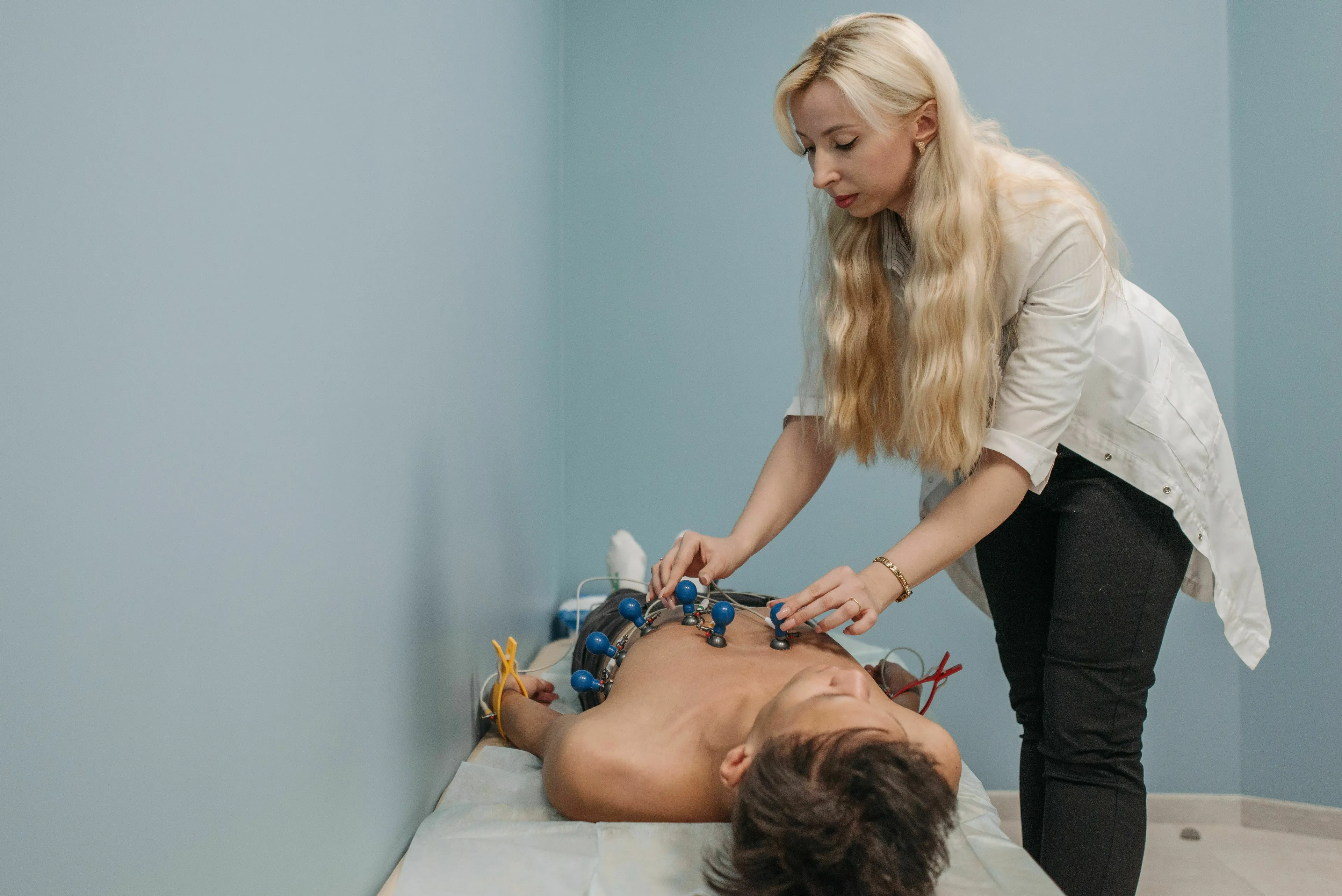 A blonde doctor in a white coat attaches electrodes to a shirtless male patient's chest during a human biology exam.