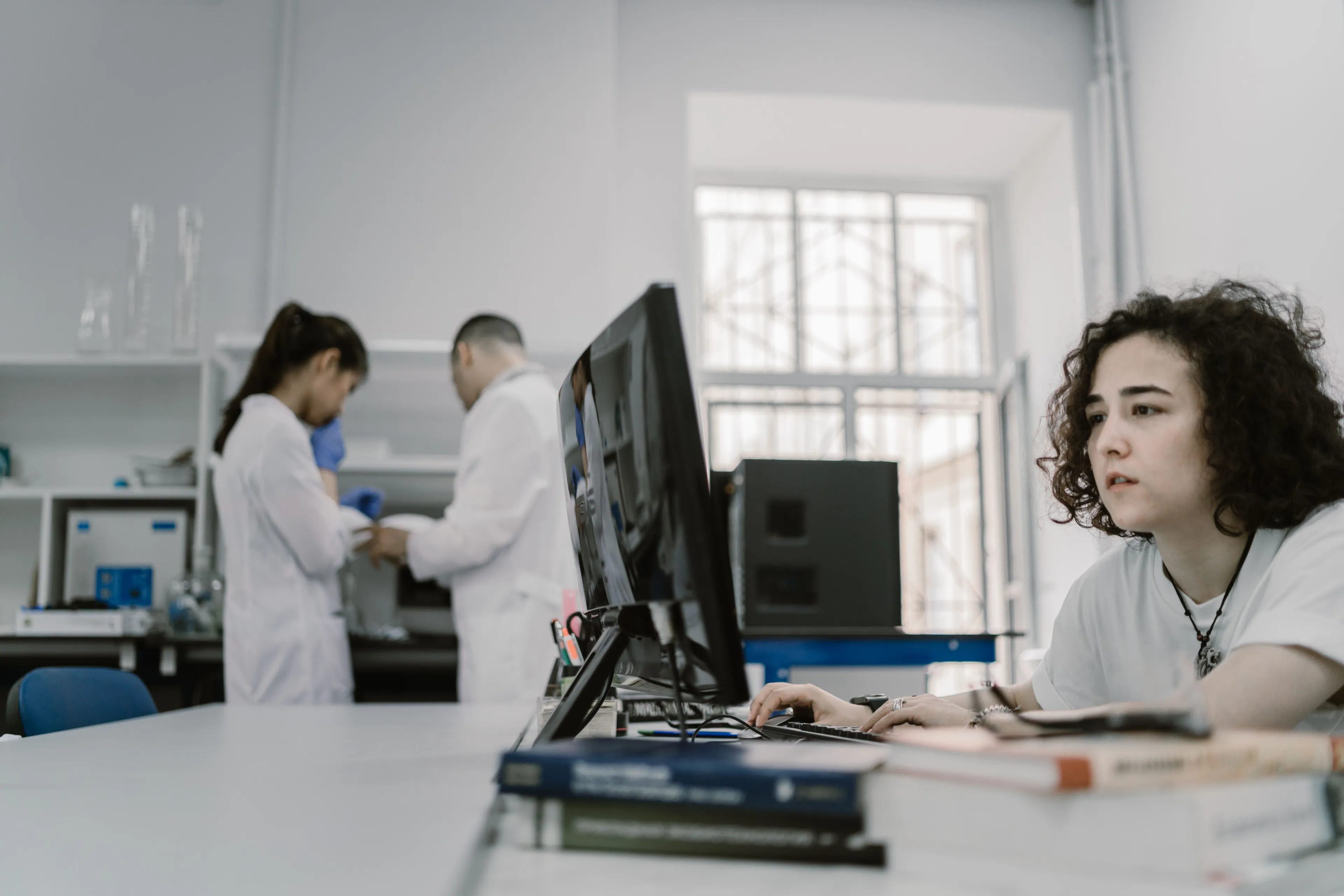 A woman with curly hair works at a computer in a science human biology lab, with two scientists in the background.