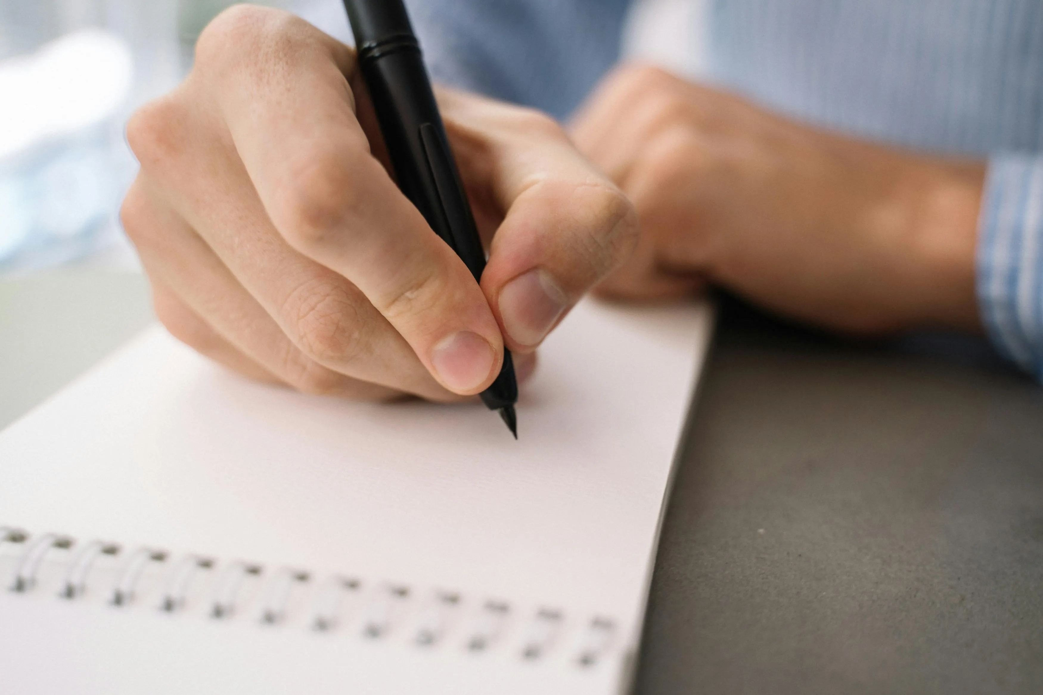 A person's hand holds a black pen, writing on a white spiral notebook, focusing on how English words are spelled.