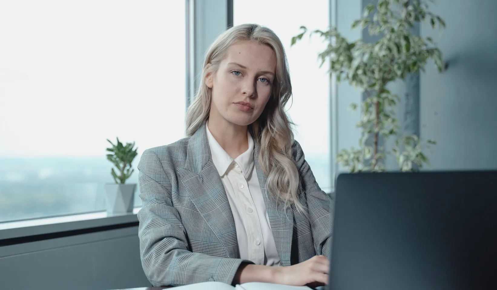 A blonde woman in a grey plaid blazer working on a laptop, contemplating how a word is spelled.