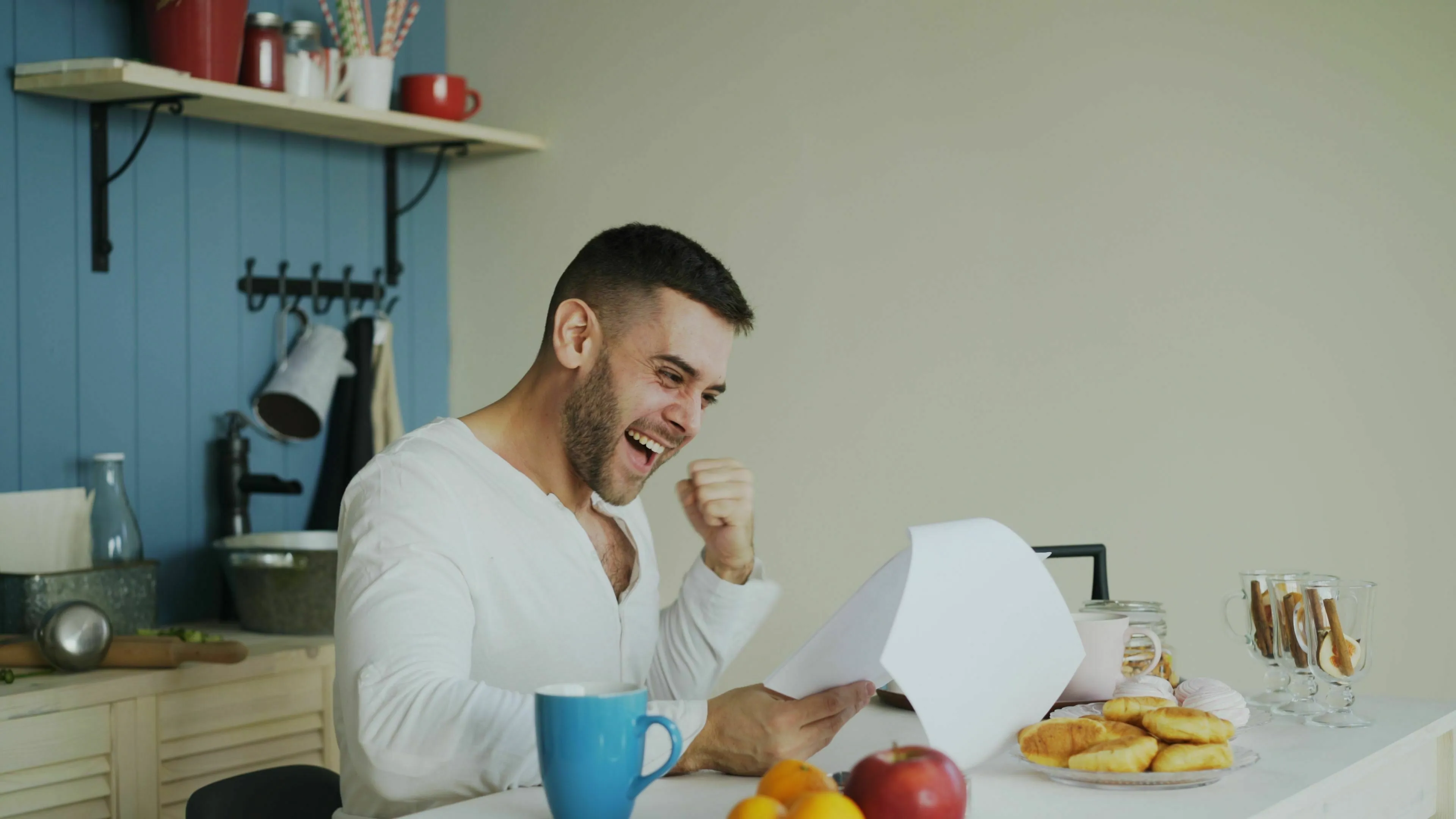 A man in a white shirt experiences joy, fist-pumping while reading good news at a kitchen counter.