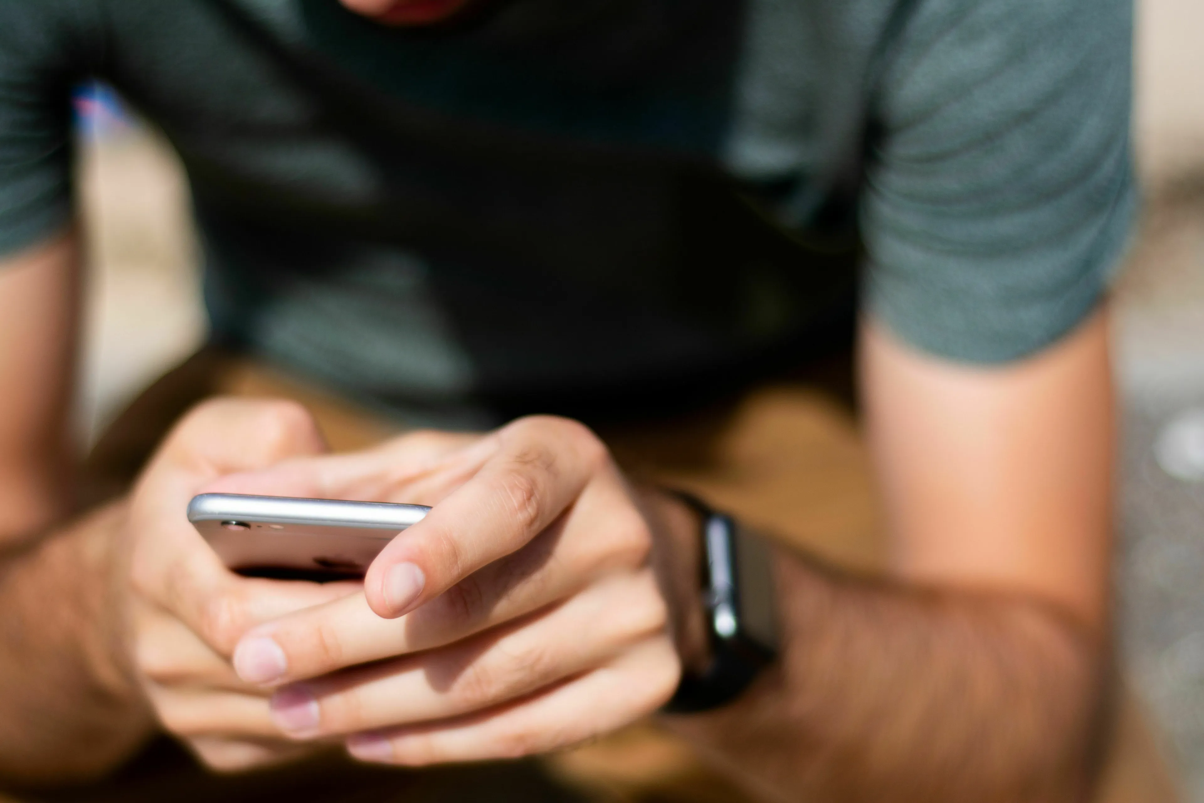 Man in a green shirt and brown pants, holding a silver smartphone. He's texting, perhaps feeling joy or disappointment.