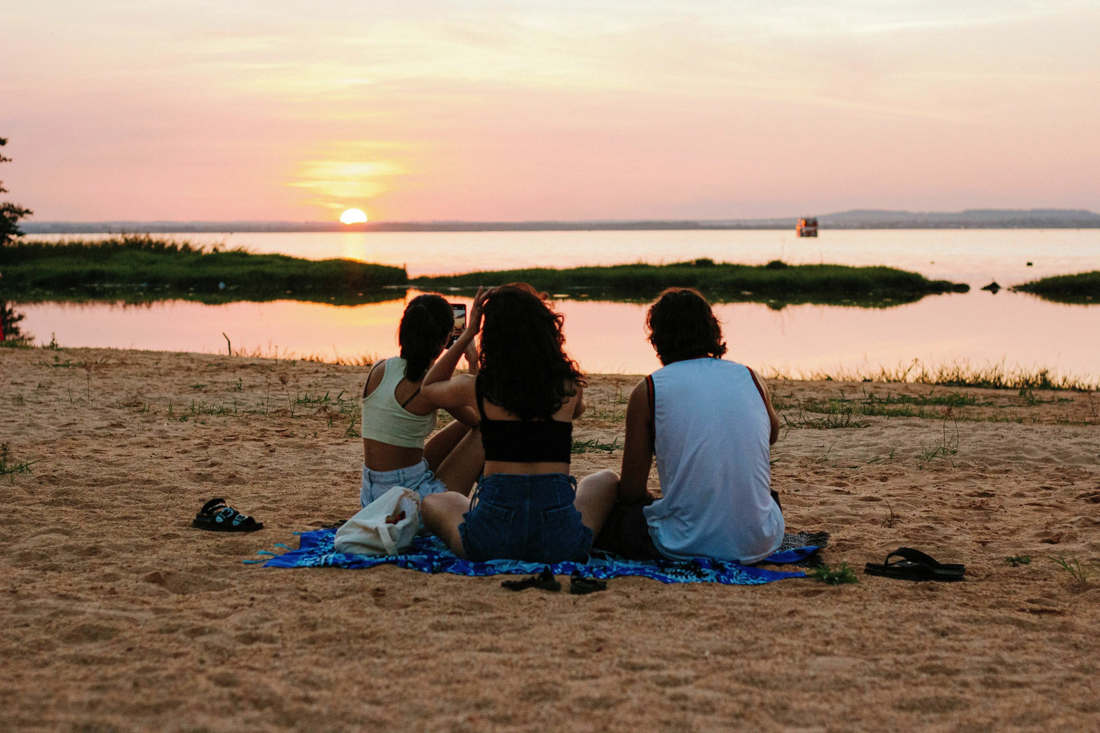 Three people sit on a beach, watching the sunset over water, experiencing joy on vacation.
