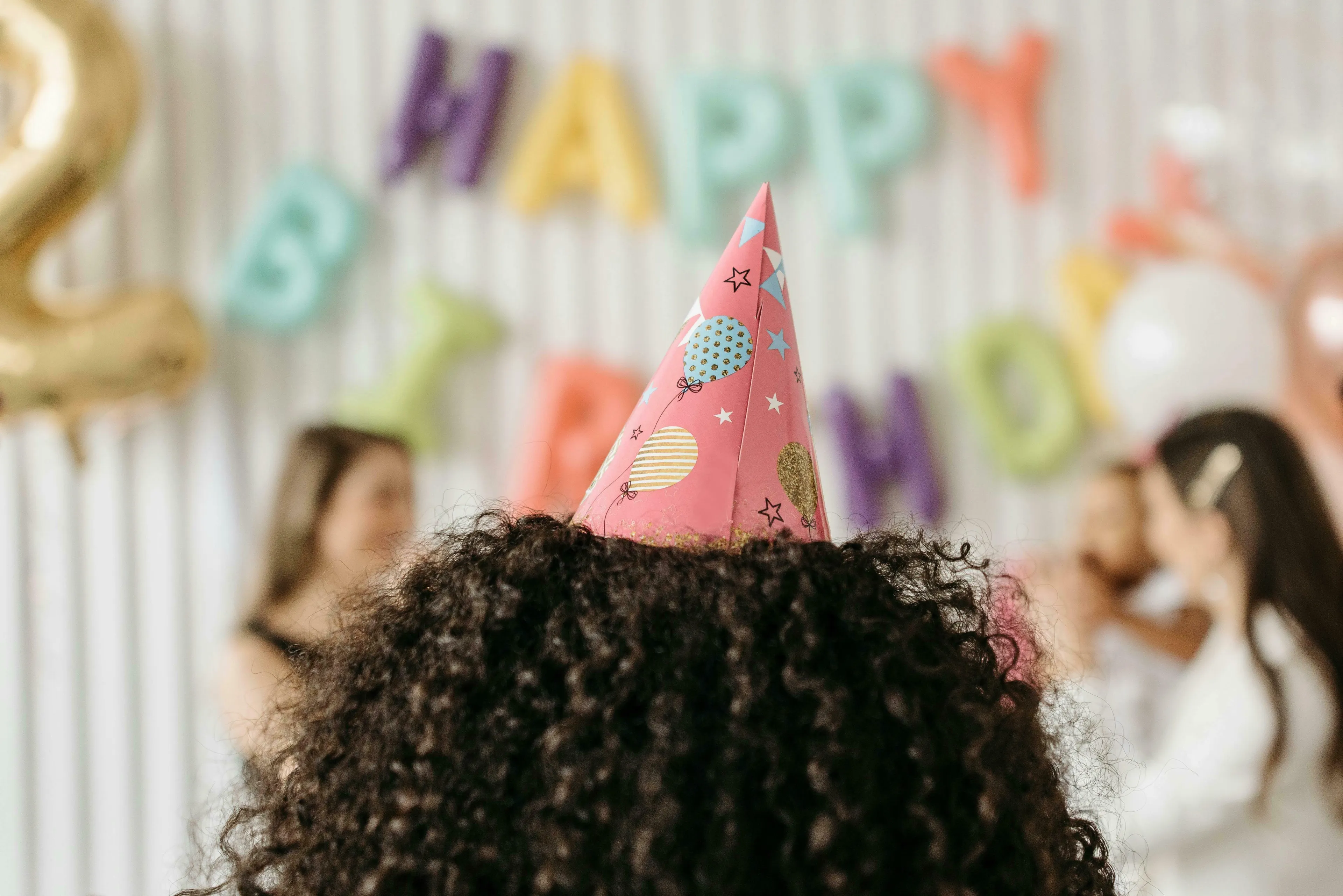 A person with curly dark hair wears a pink birthday hat, celebrating a birthday. Experience joy at the party.