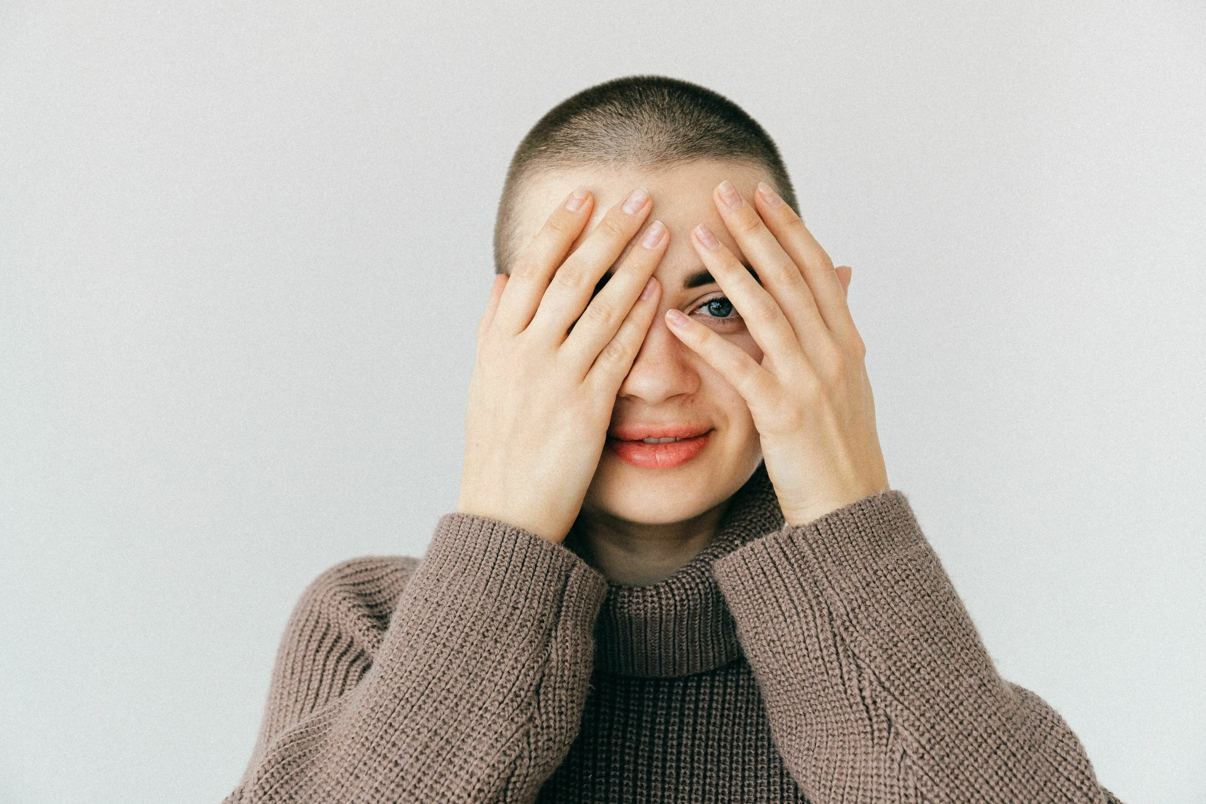 A person with a buzz cut covering their face with their hands, peeking through their fingers. Experience joy from a compliment.