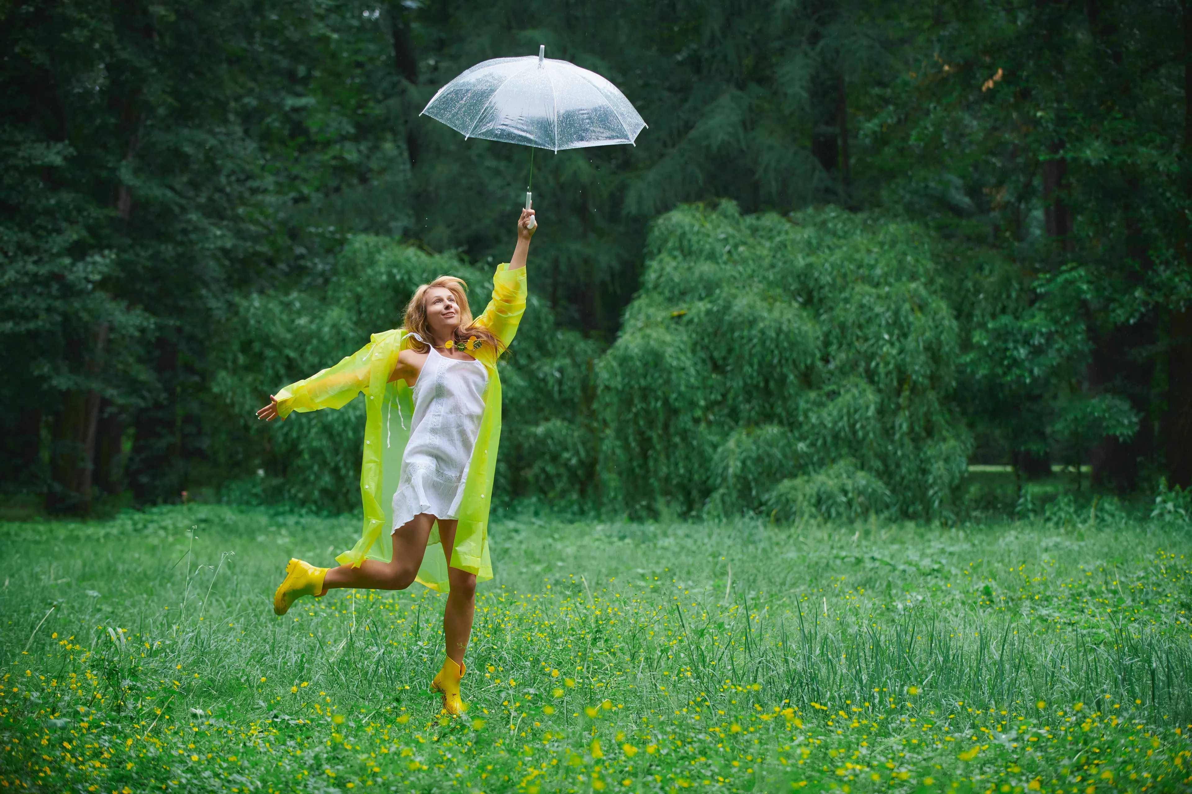 A woman in a yellow raincoat and boots leaps joyfully with an umbrella in a grassy field. Experience joy on a rainy day!