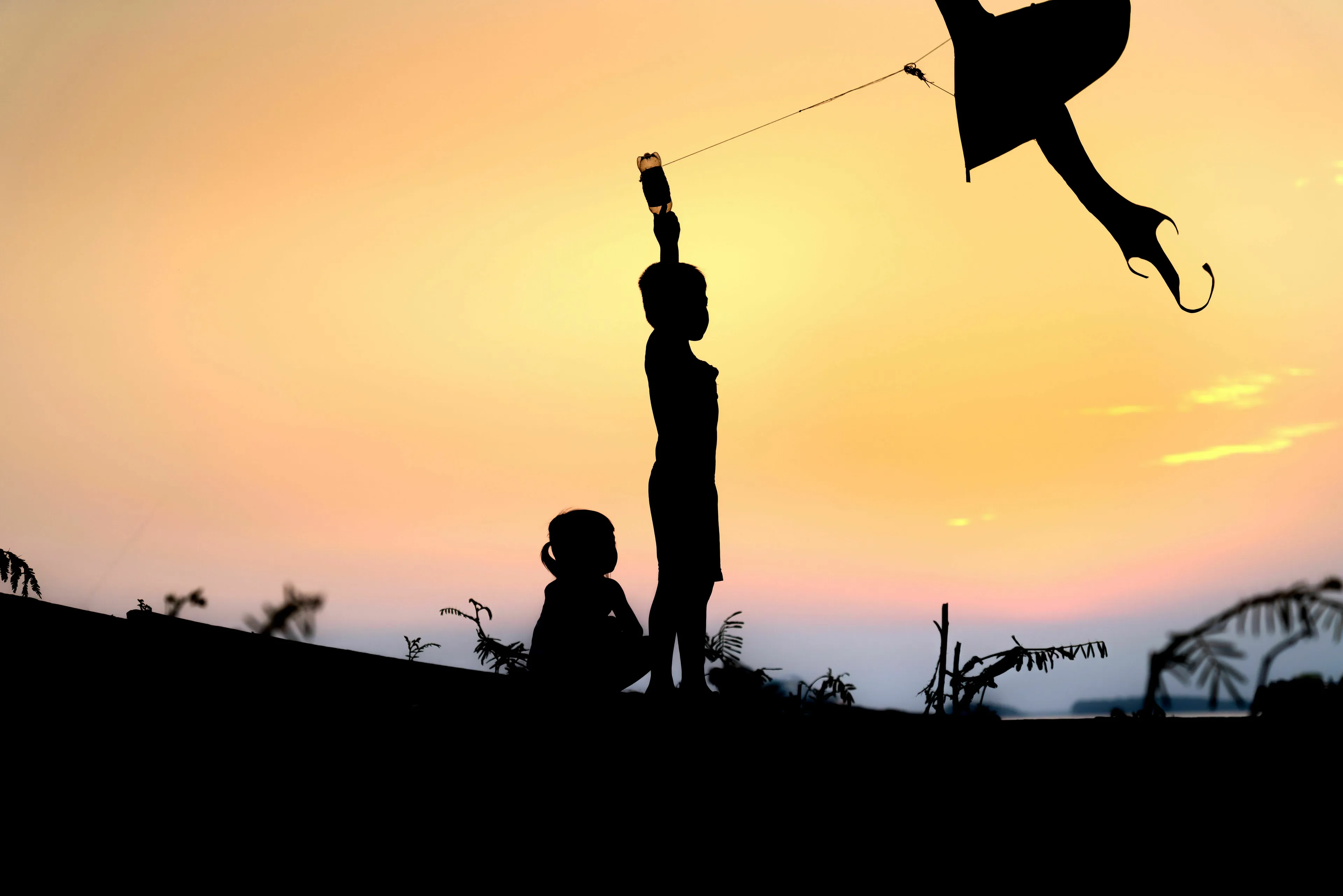Silhouette of two children flying a kite against a sunset sky, evoking joy and what makes you nostalgic.