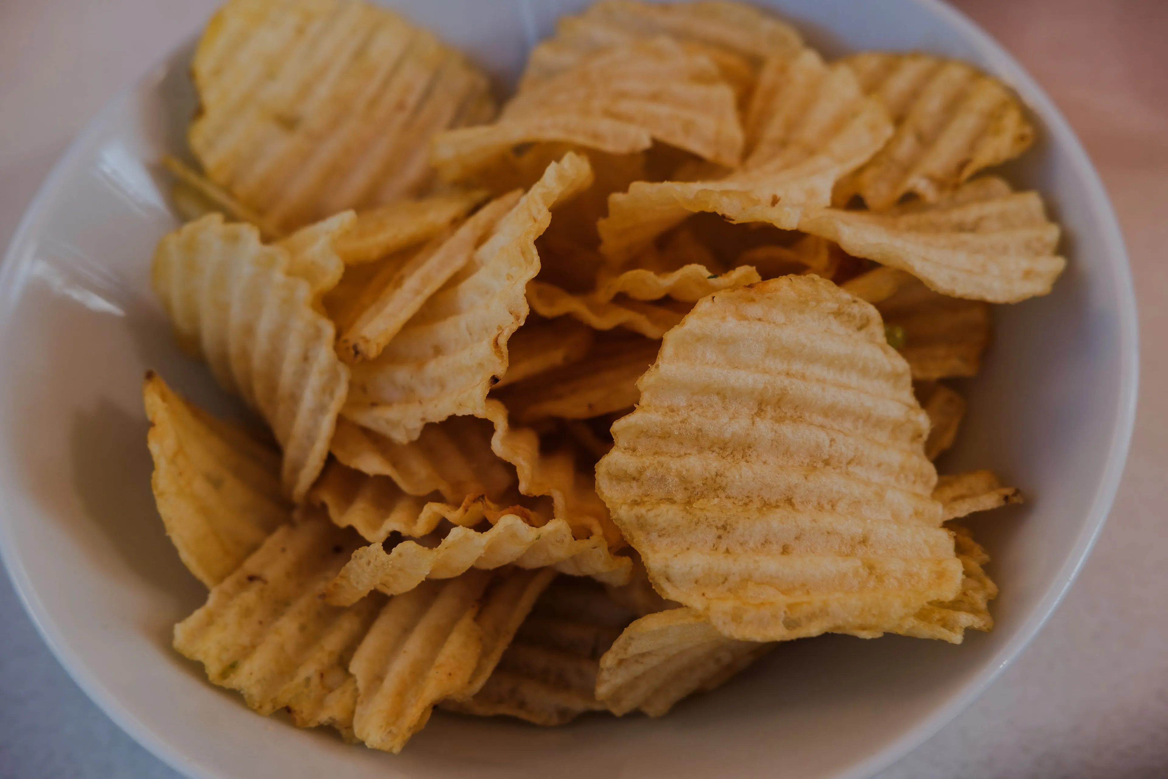 A white bowl filled to the brim with crispy, golden-yellow ripple cut chips, ready to be a delicious snack.