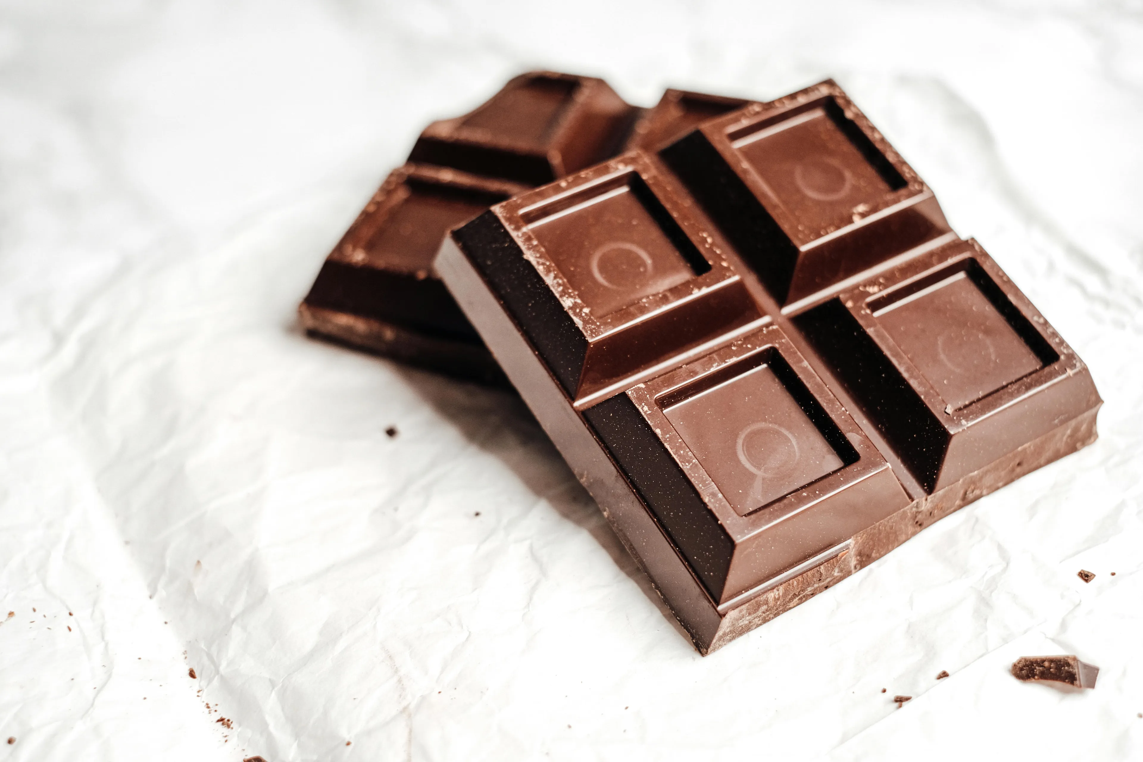 Two dark chocolate bars, partially broken, sit on crinkled white paper, a tempting snack option.