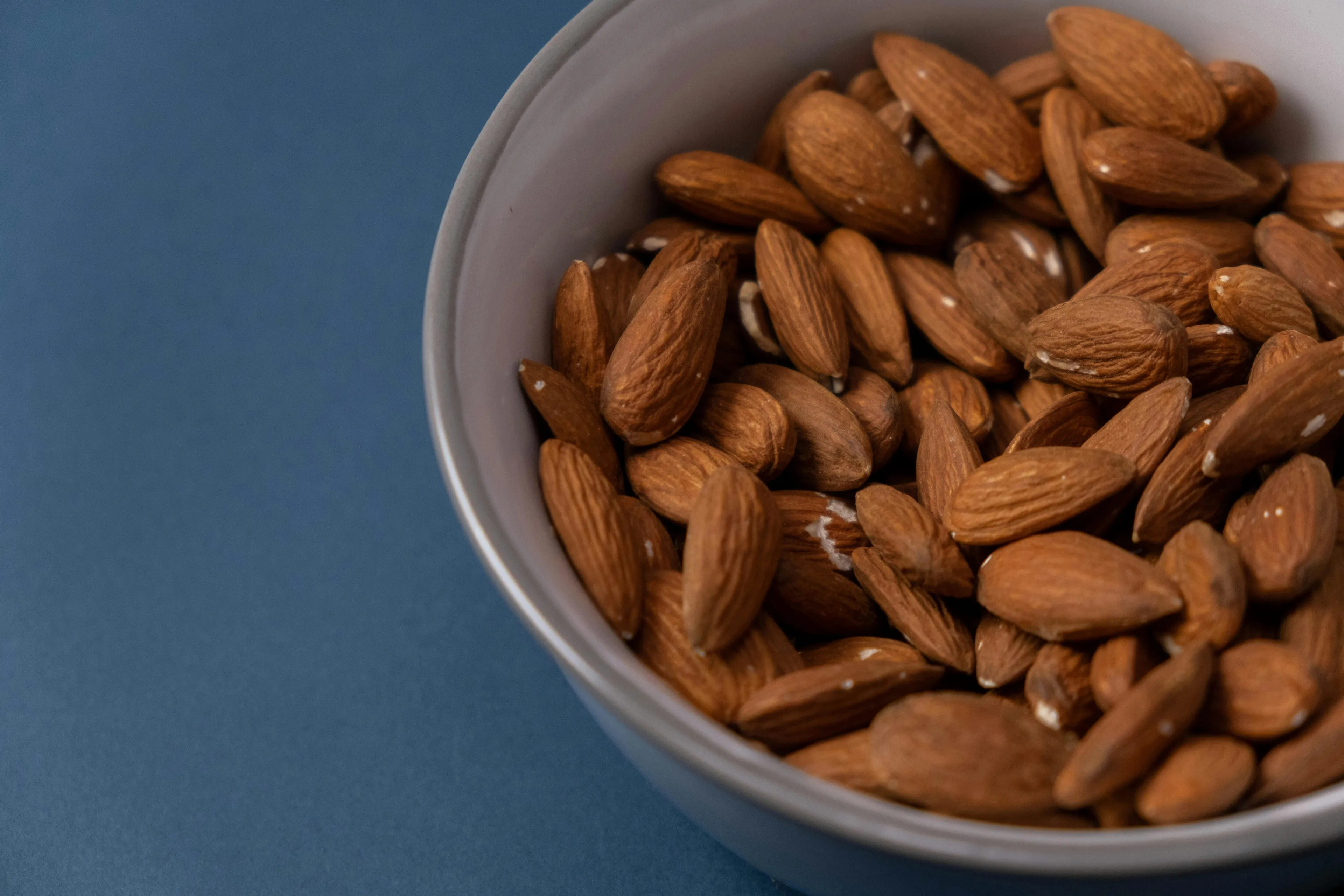 A white bowl filled with raw almonds, ready to pick a snack, resting on a dark blue surface.