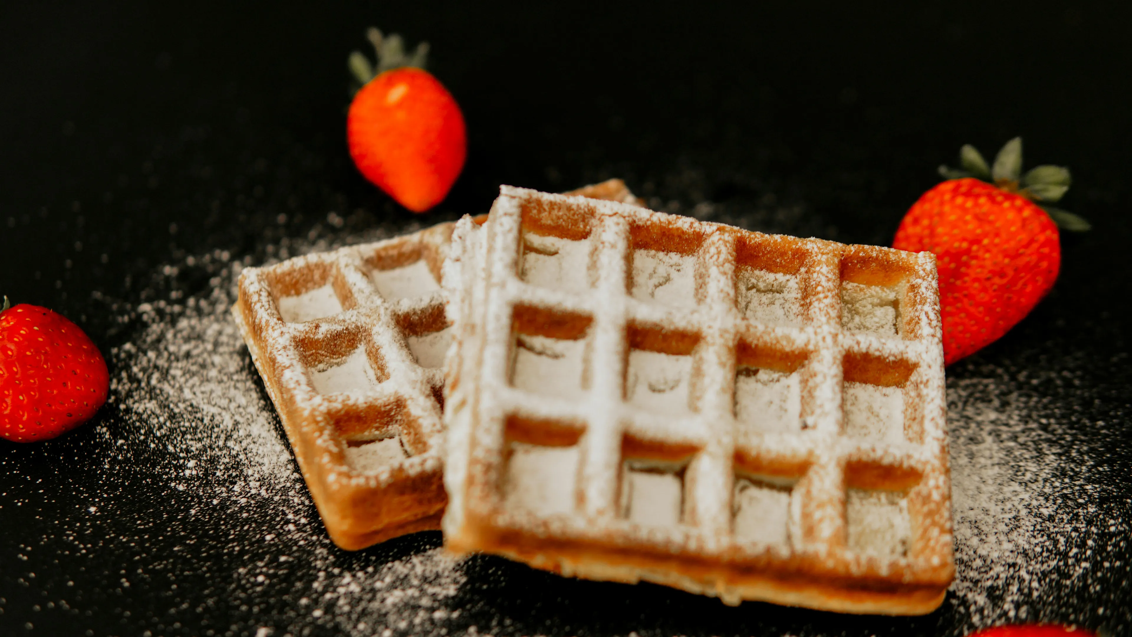 Delicious waffles dusted with powdered sugar, surrounded by fresh strawberries on a dark surface. A perfect snack!