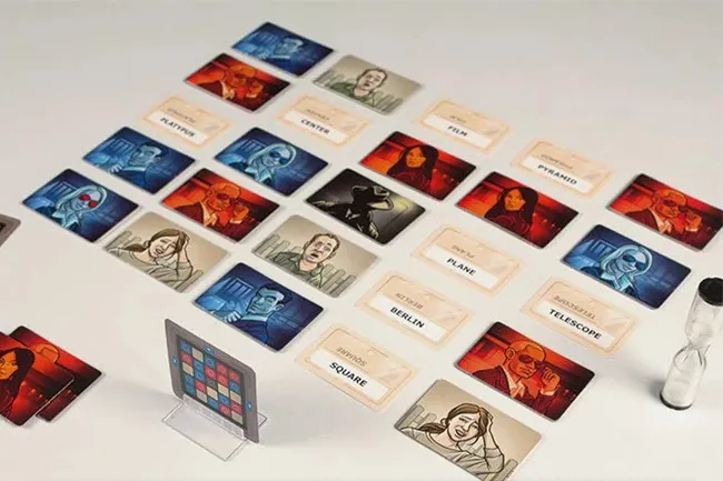 A spread of Codenames board game cards on a white surface, featuring clue words and agent illustrations, with an hourglass nearby.