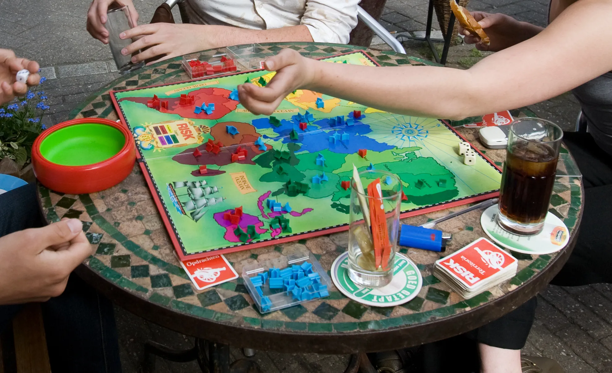 Friends playing Risk board game outdoors, with drinks on a mosaic table. A classic board game choice.