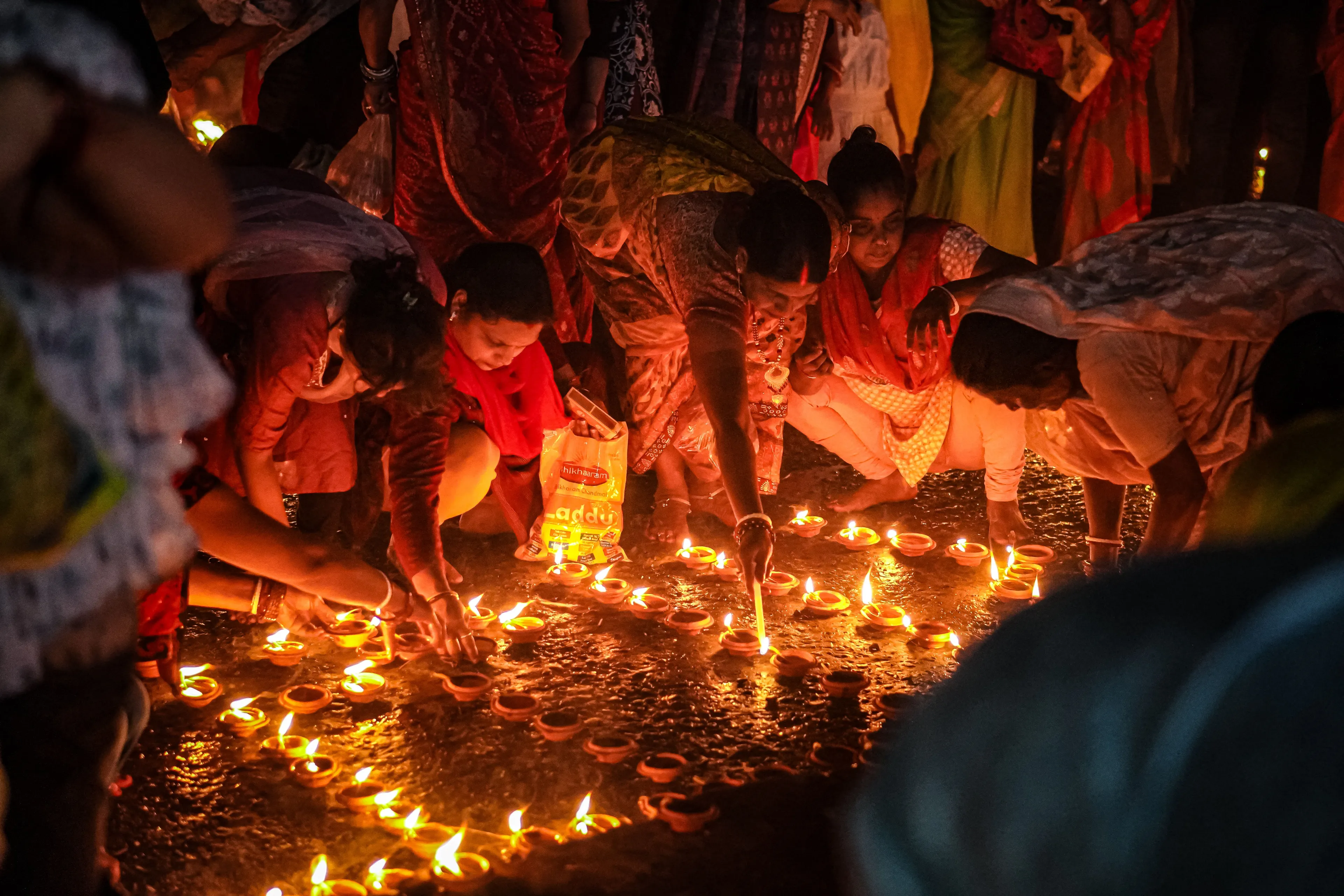 People lighting numerous small oil lamps during the Diwali festival, illuminating a dark, wet ground.