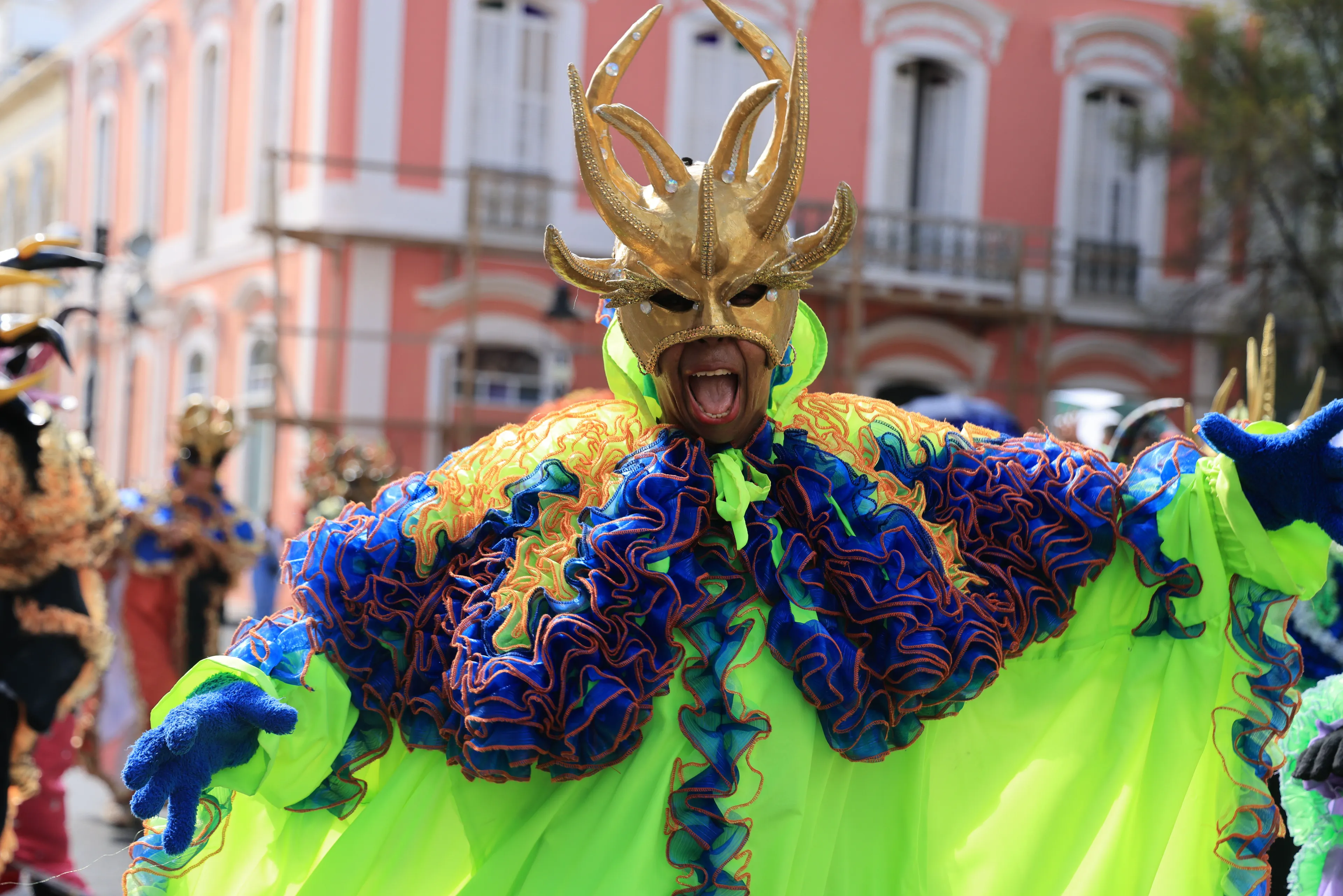 A vibrant performer in a golden mask and elaborate neon green and blue costume shouts joyfully during Rio Carnival.