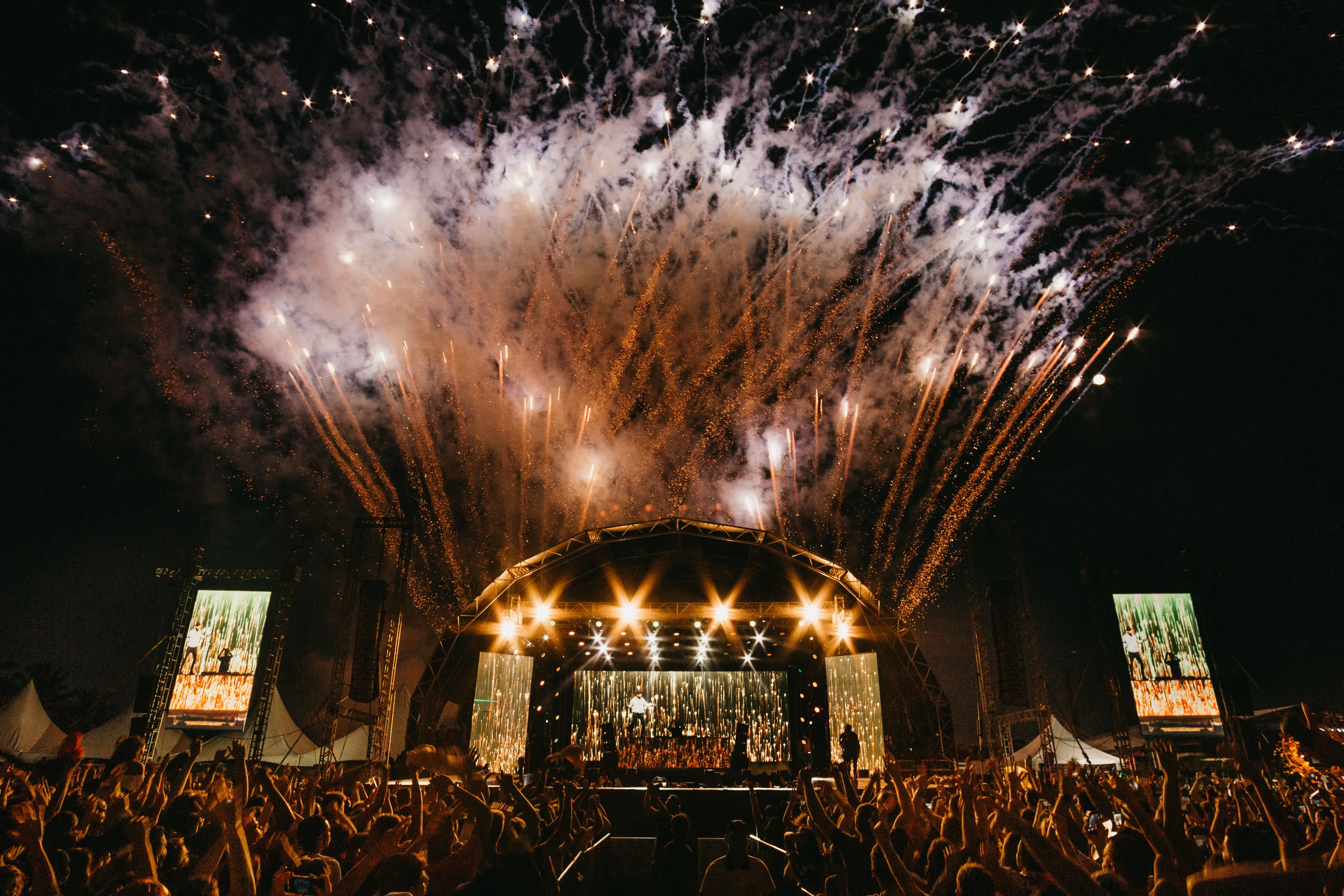 Fireworks explode above a crowded outdoor concert stage at night, a famous festival like Coachella.