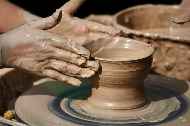 Hands covered in clay shaping a vessel on a pottery wheel, illustrating the craft of pottery.