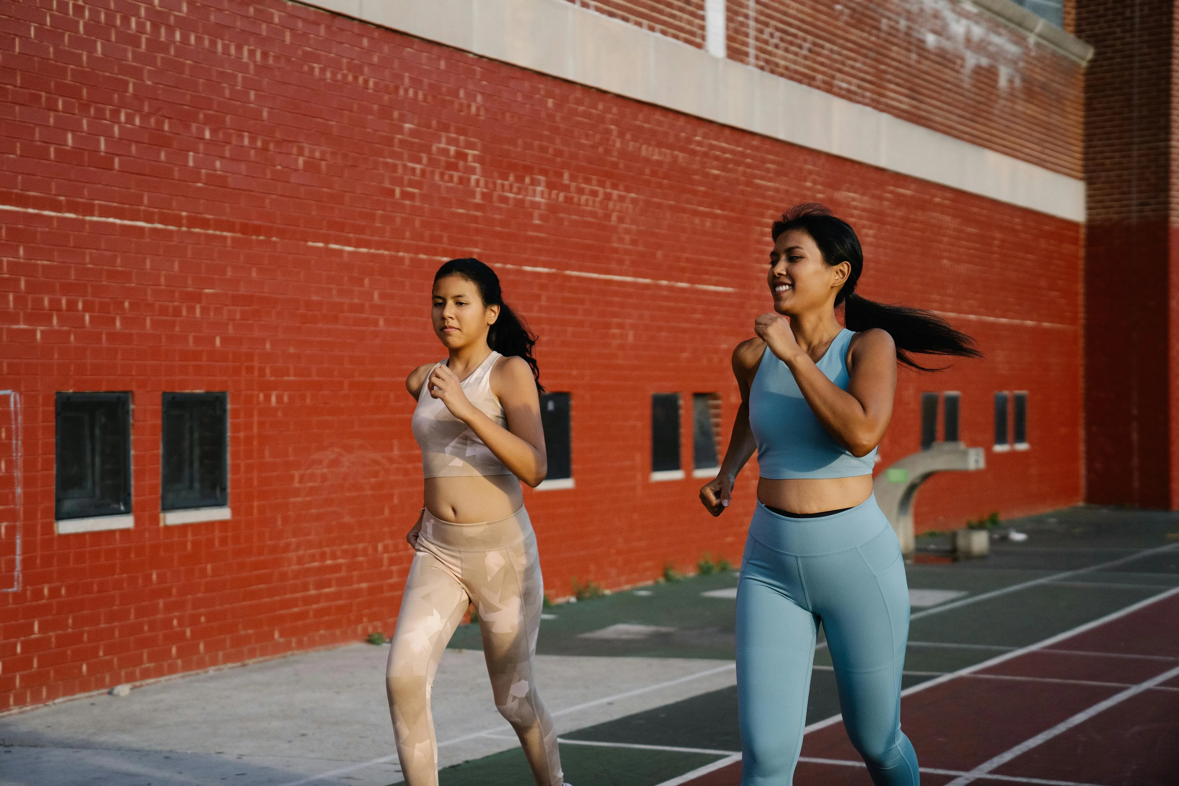 Two women in athletic wear, one in beige and one in blue, smiling while running on a track.
