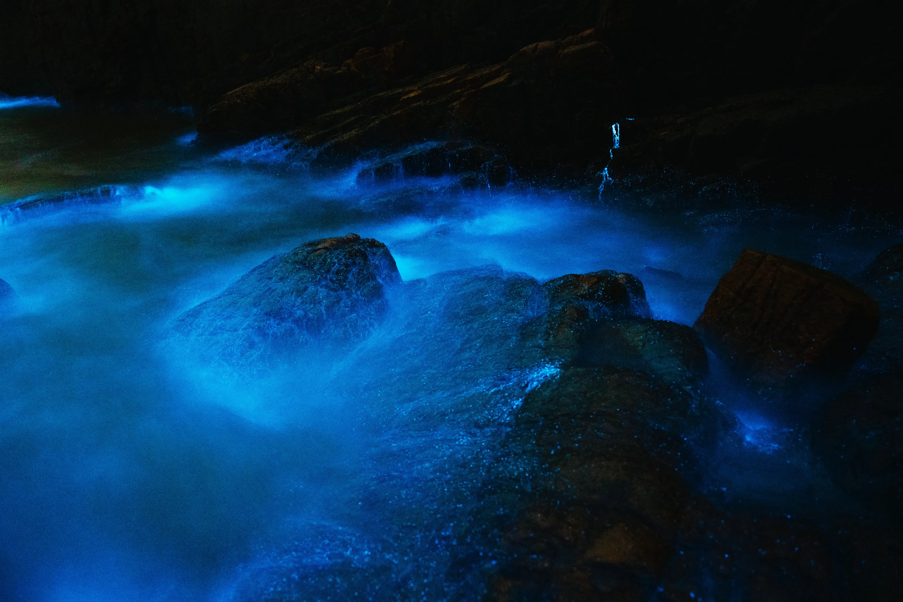 Stunning bioluminescence in a dark grotto, bright blue light illuminating water flowing over dark rocks and through caves.