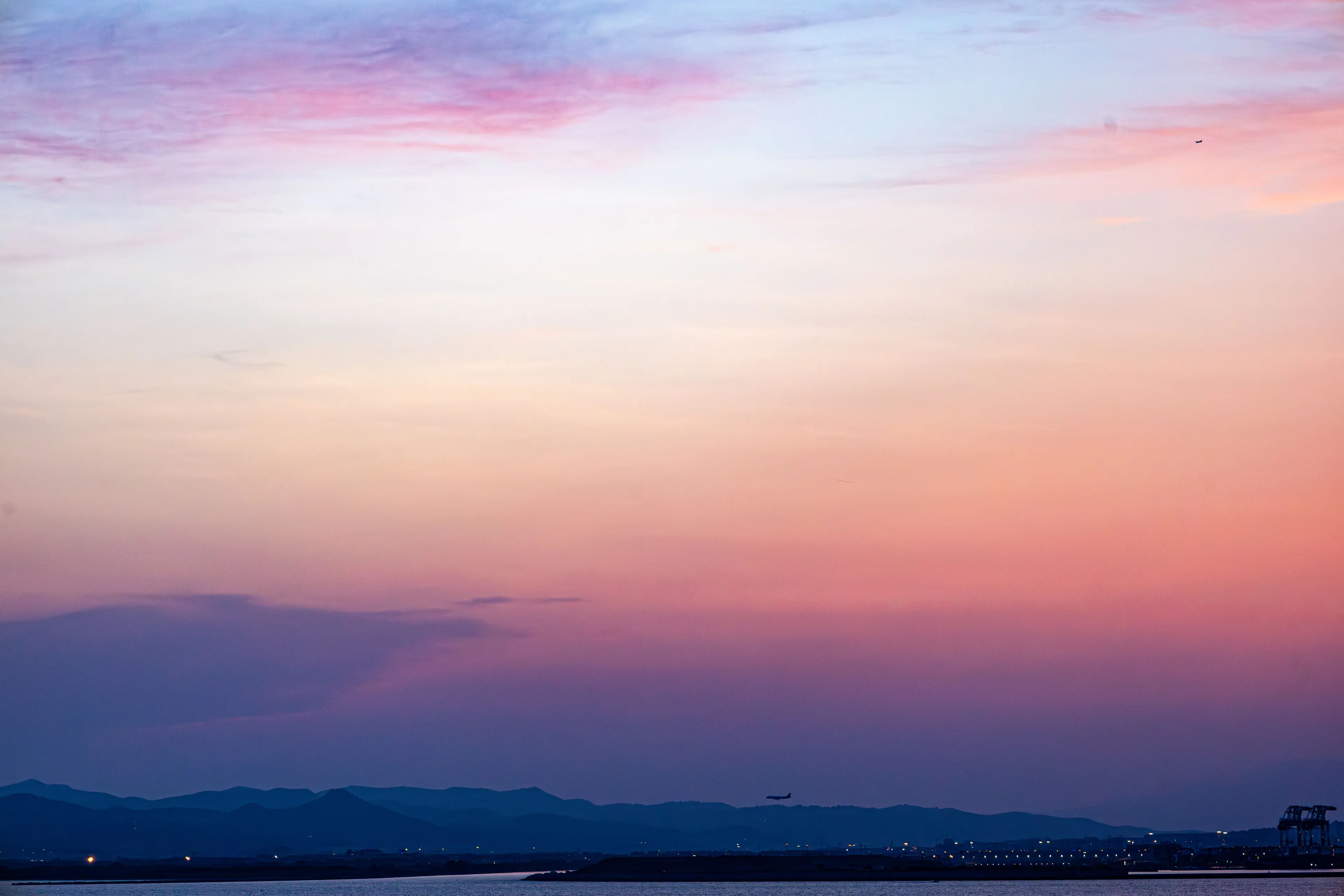 A beautiful sunrise with a sky showcasing a type of light that transitions from soft blues to vibrant pinks and deep purples. A plane flies above mountains.