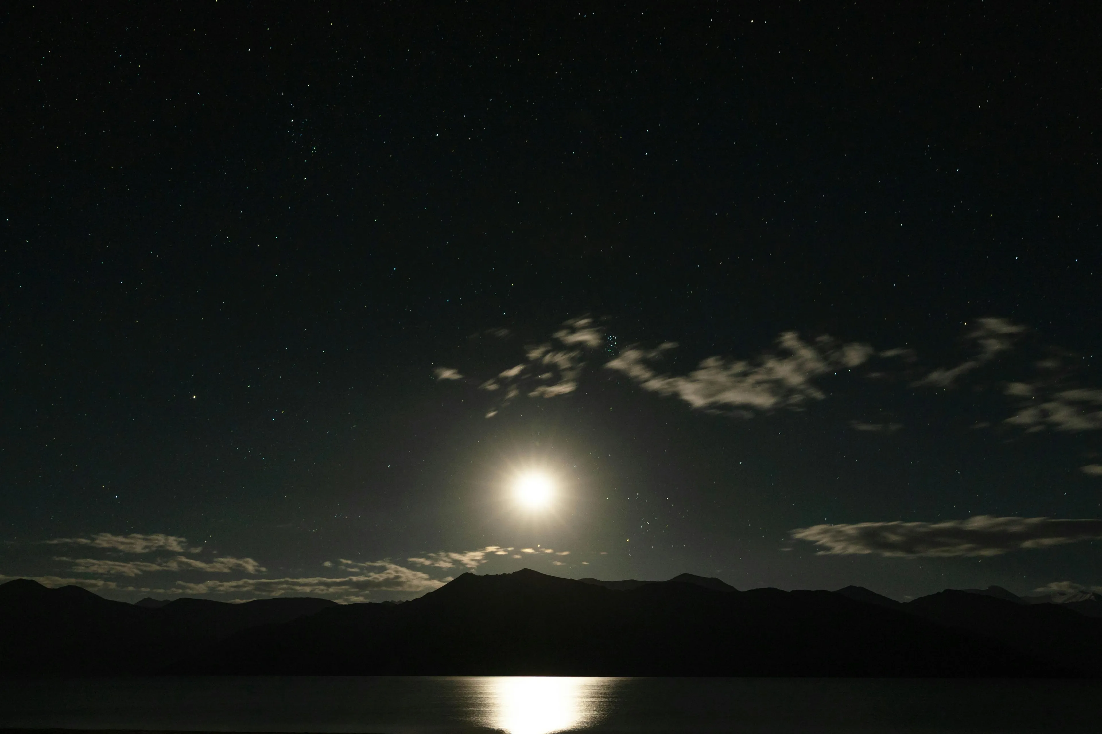 Moonlight reflecting on a lake at night, silhouetted mountains against a star-filled sky with scattered clouds.