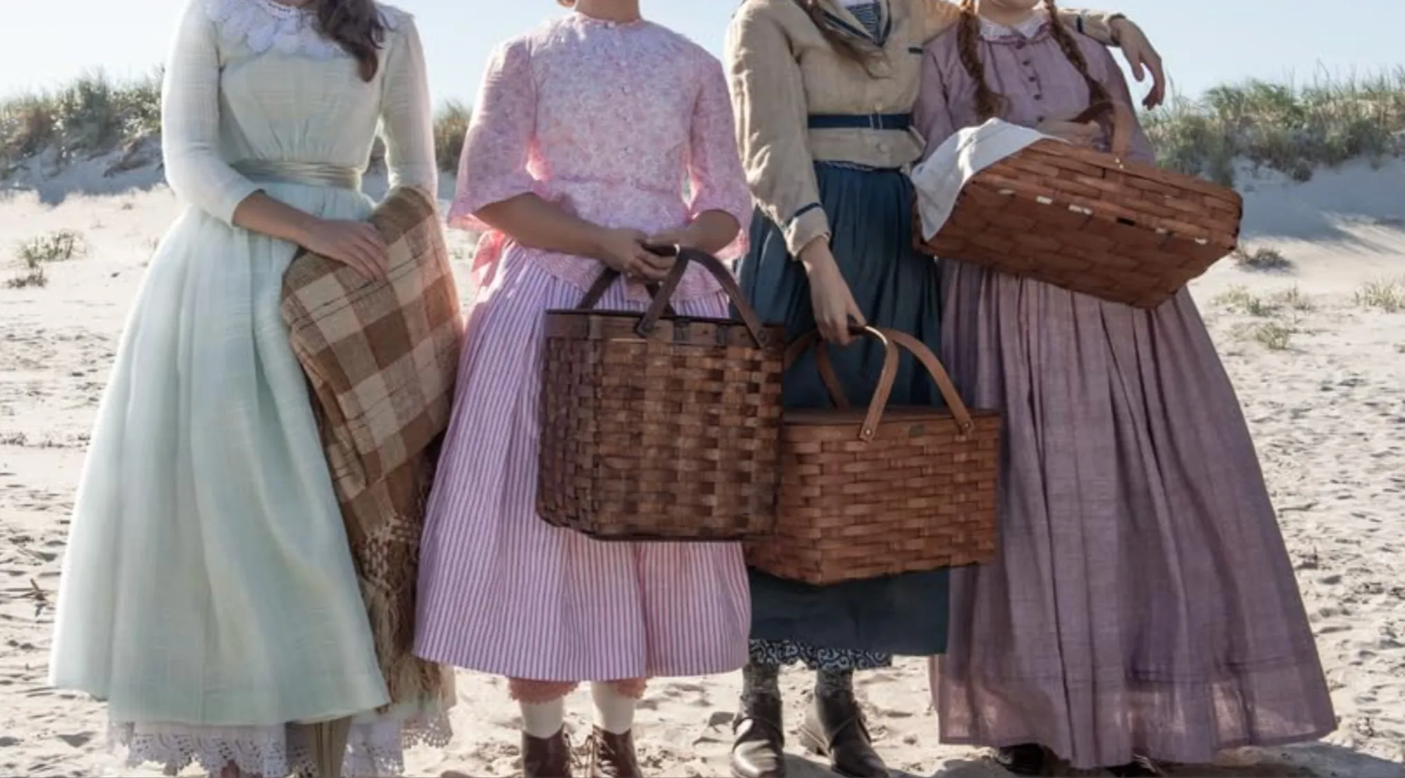 Four young women in iconic outfits from Little Women carrying picnic baskets on a sandy beach.