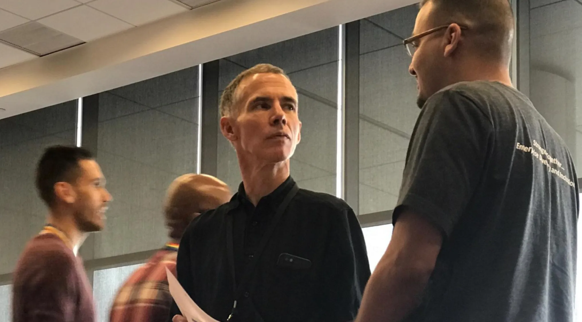 A man in a black shirt converses with another man; potential celebrity doppelganger.