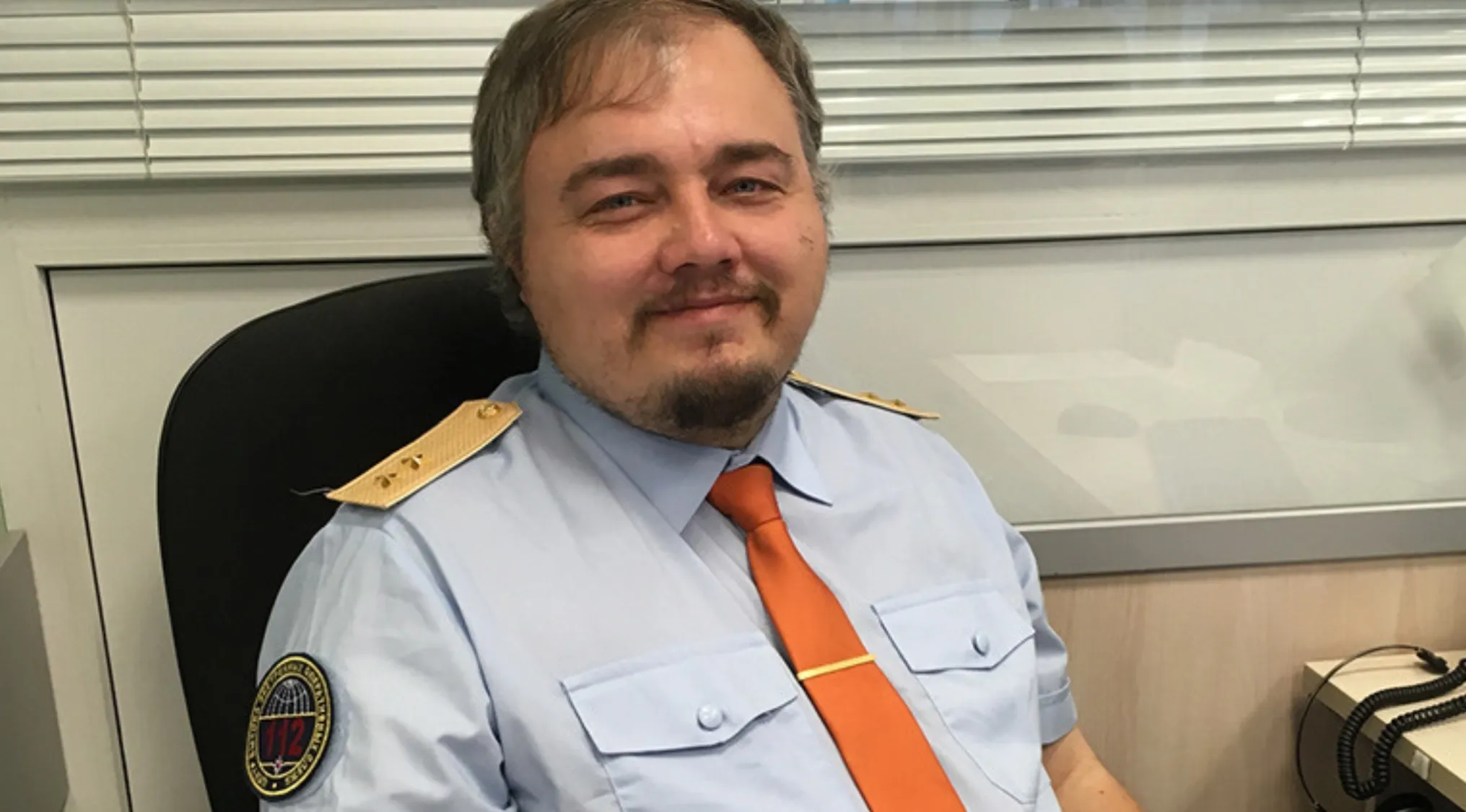 Smiling man in a light blue uniform shirt with an orange tie. A celebrity doppelganger. Who does this person remind you of?