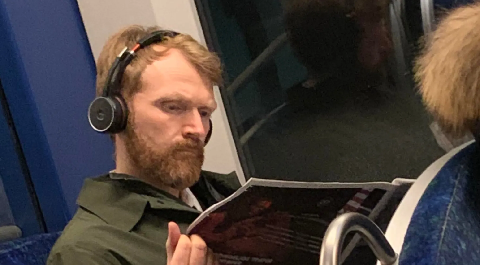 A red-haired man with a beard and headphones reads a magazine on a train. Does he have celebrity doppelgangers? Which painter does he look like?