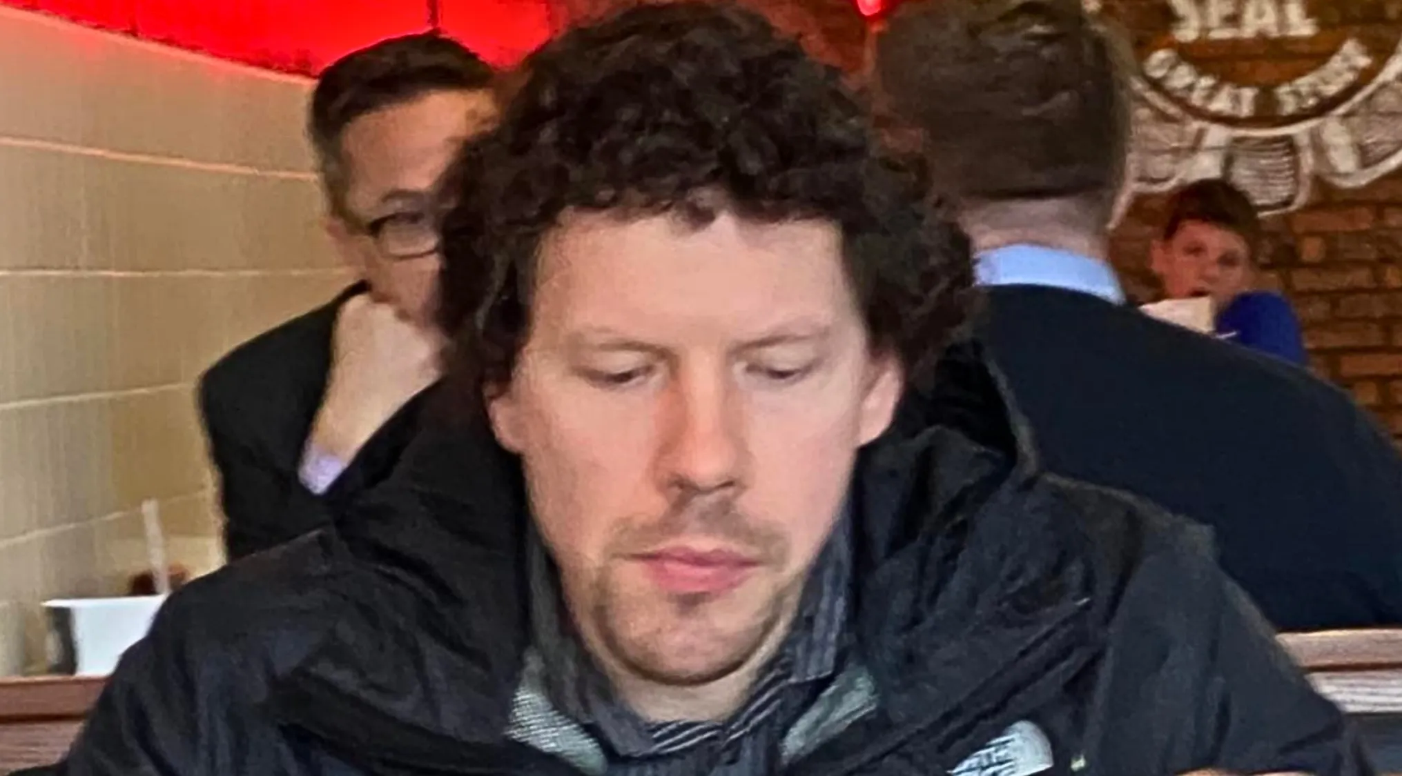 Man with curly hair and a beard, wearing a black jacket, looking down. A celebrity doppelganger.