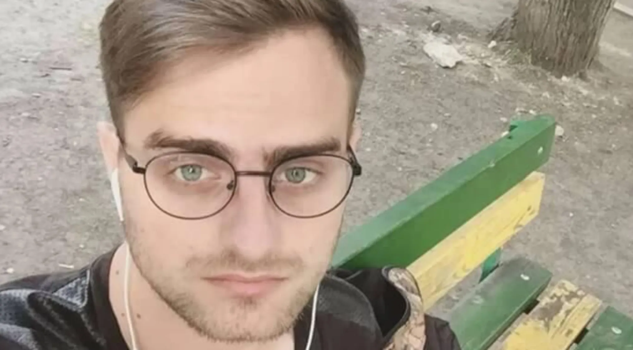 A young man with glasses and earbuds, looking serious, seated on a green and yellow bench. Is he a celebrity doppelganger?