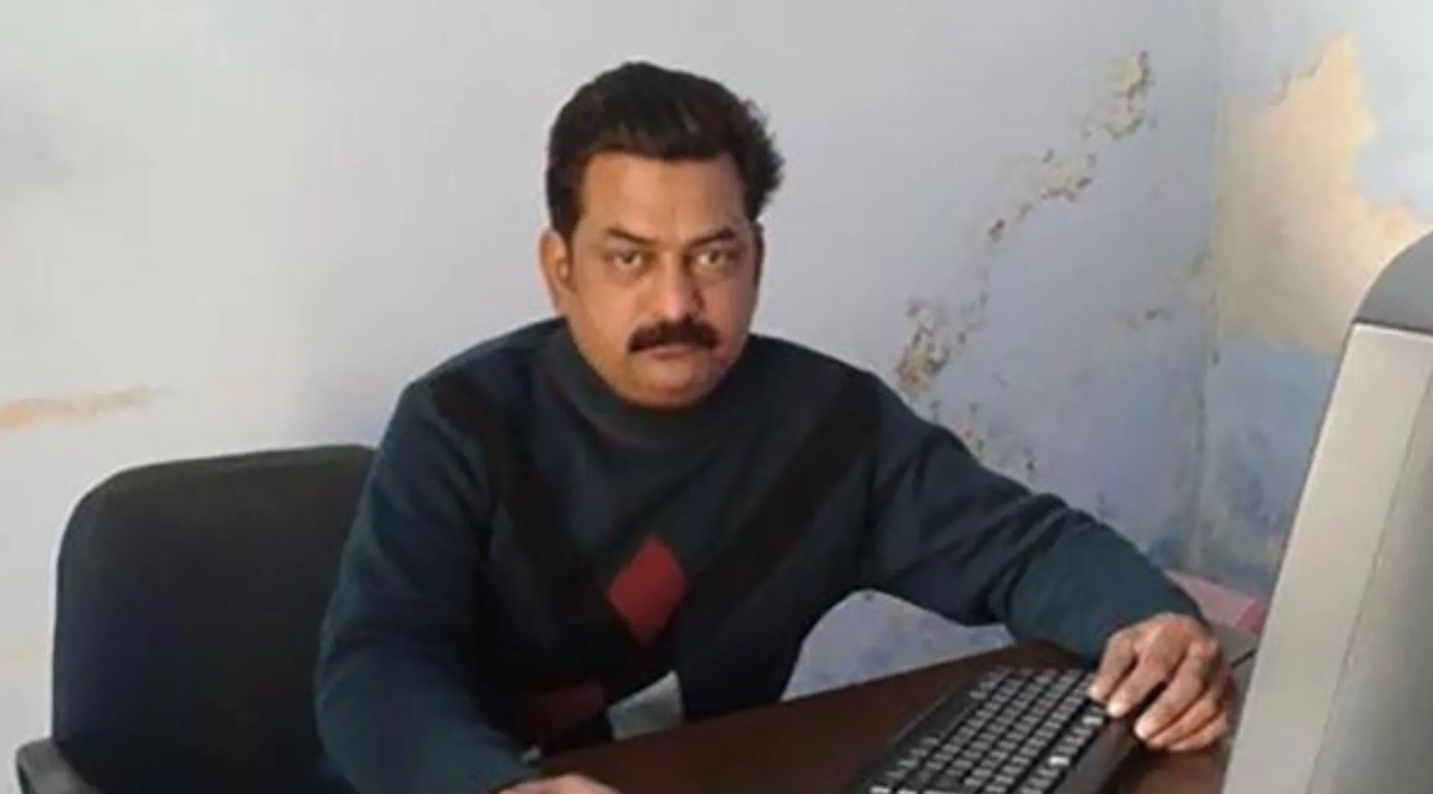 A man with a mustache, dark hair, and a dark argyle sweater sits at a desk, looking like celebrity doppelgangers.
