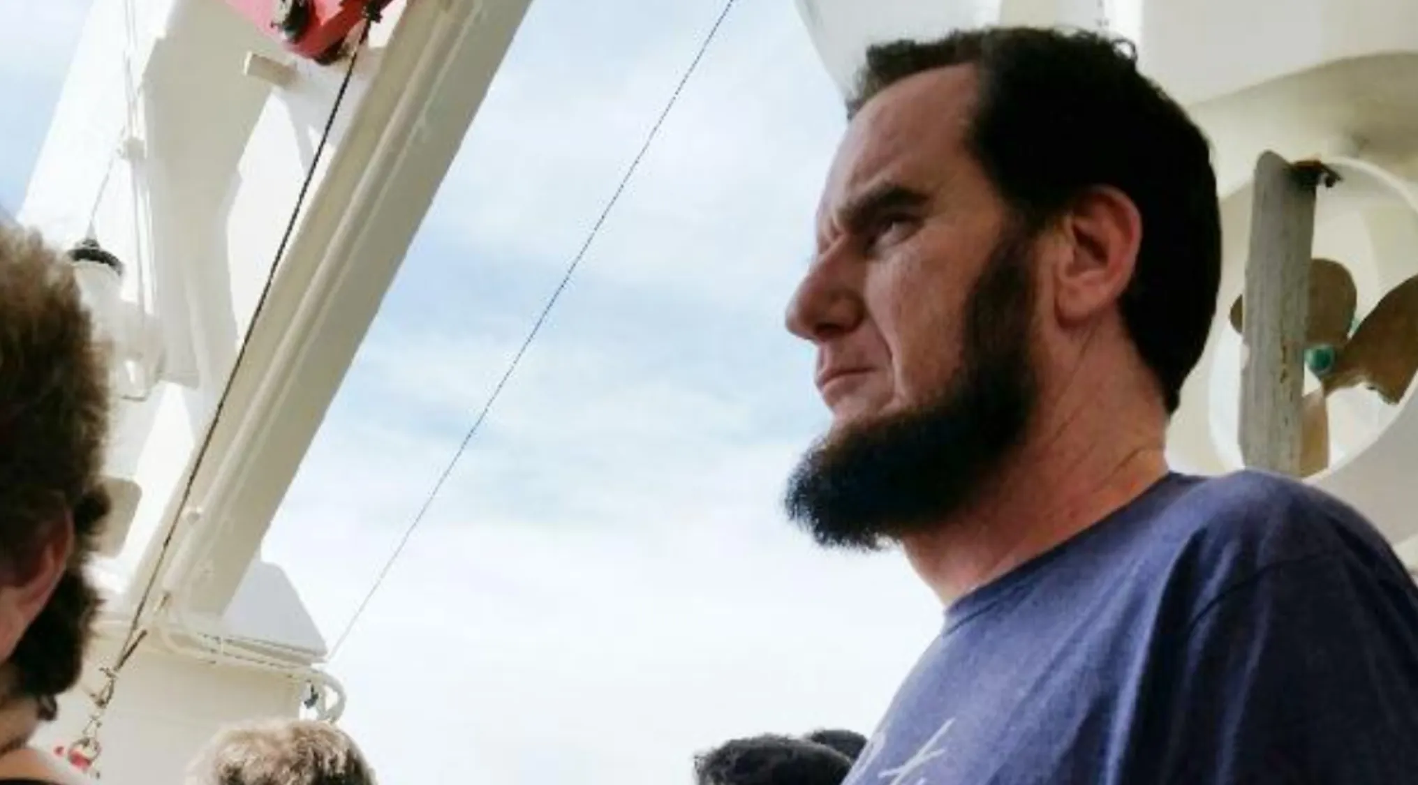 A man with a beard and dark hair, resembling a Celebrity Doppelganger, looks right. He wears a blue shirt against a bright sky.
