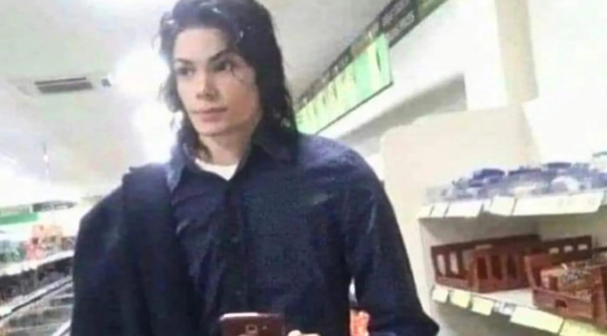 A person with long dark hair, resembling Michael Jackson, holding a phone in a store aisle. Celebrity doppelgangers.