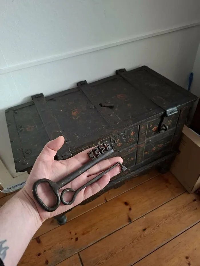 A hand holding large, antique iron keys in front of a rustic wooden chest filled with old treasures.