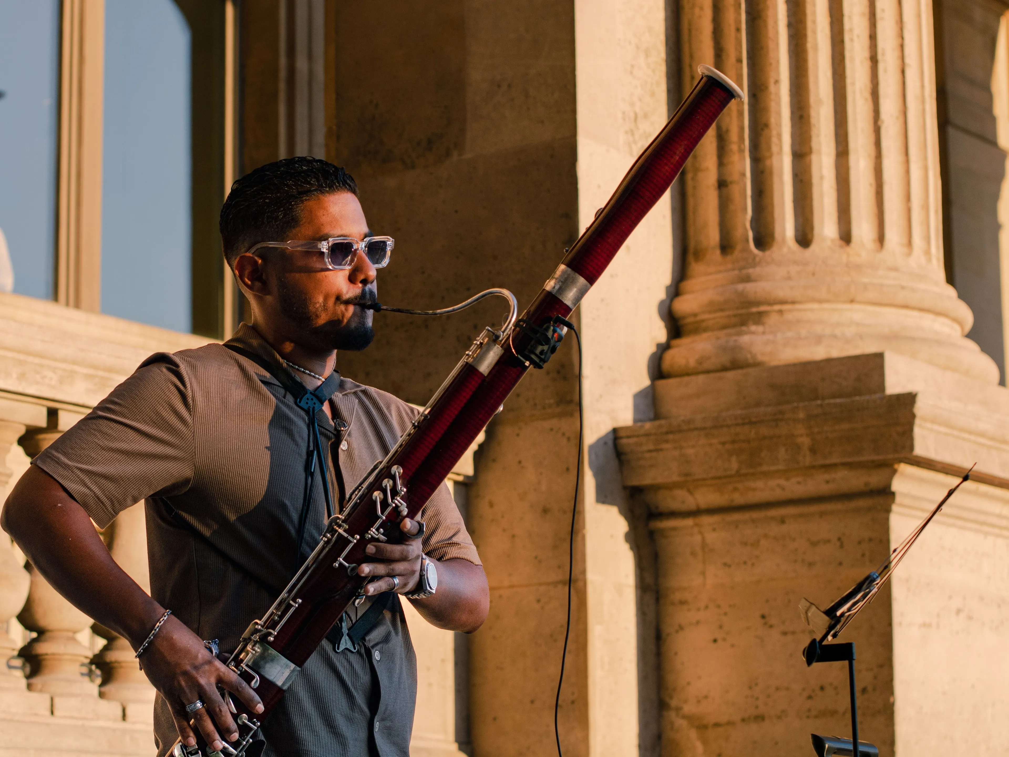 A man wearing sunglasses and a brown shirt playing the bassoon, with classical Art columns in the background.