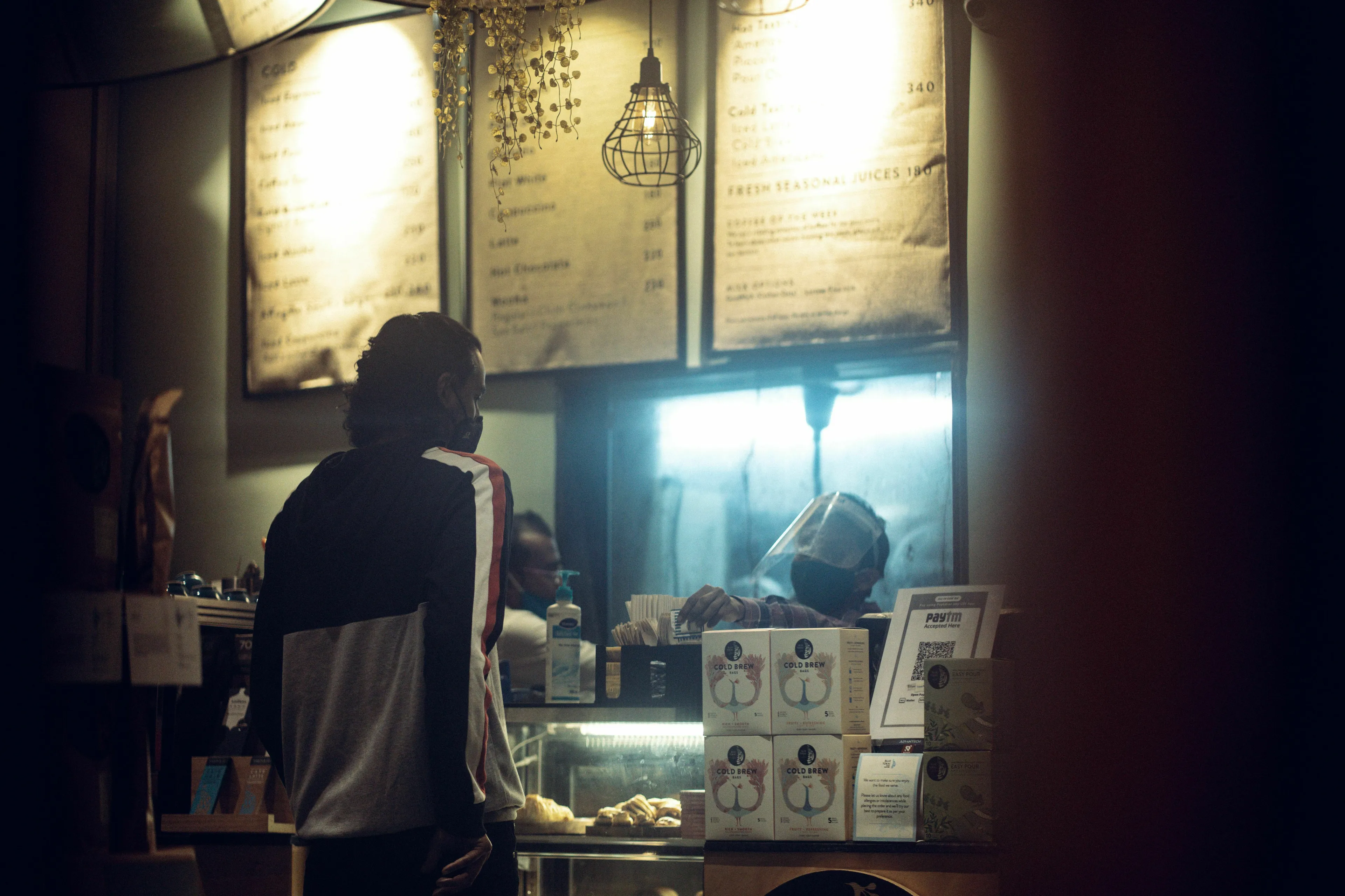 A person wearing a mask stands at a coffee shop counter. Are you extroverted? Where do you sit in a coffee shop?
