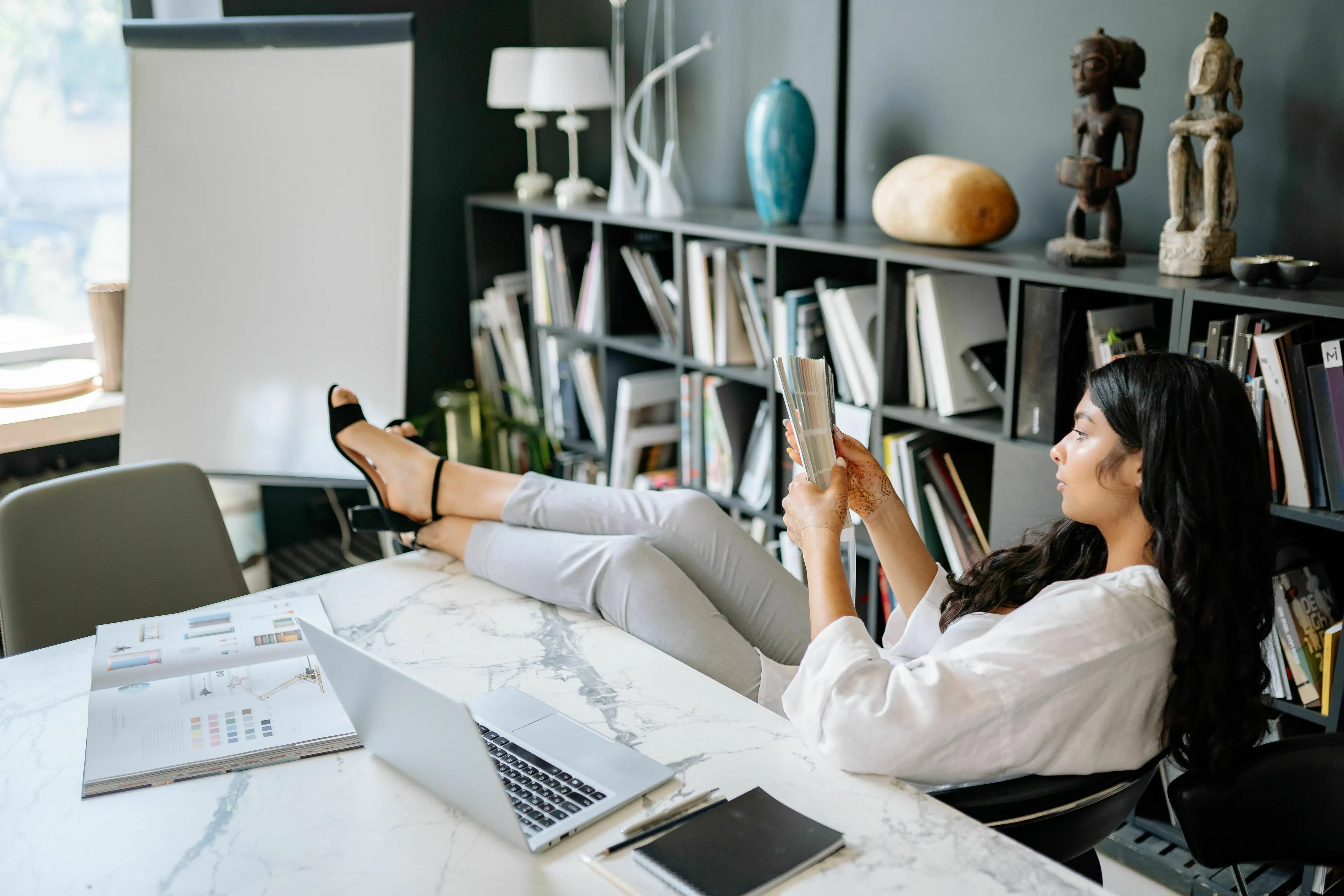 A woman relaxing with her feet on a desk, reading, considering her ideal workday and how extroverted she is.