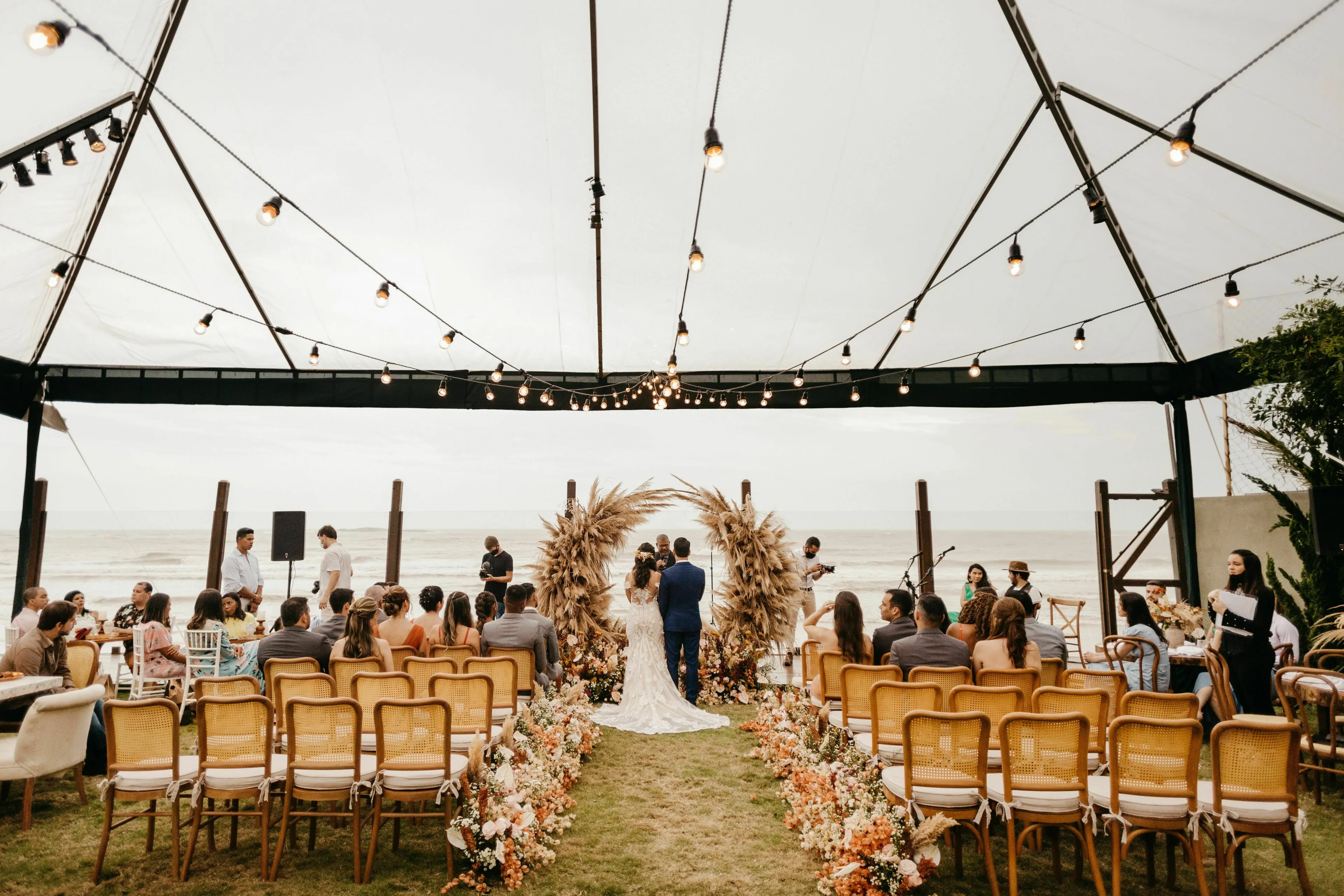 A couple weds on a beach under a tent with string lights. Guests are seated in rows. Where will you be found at this wedding?