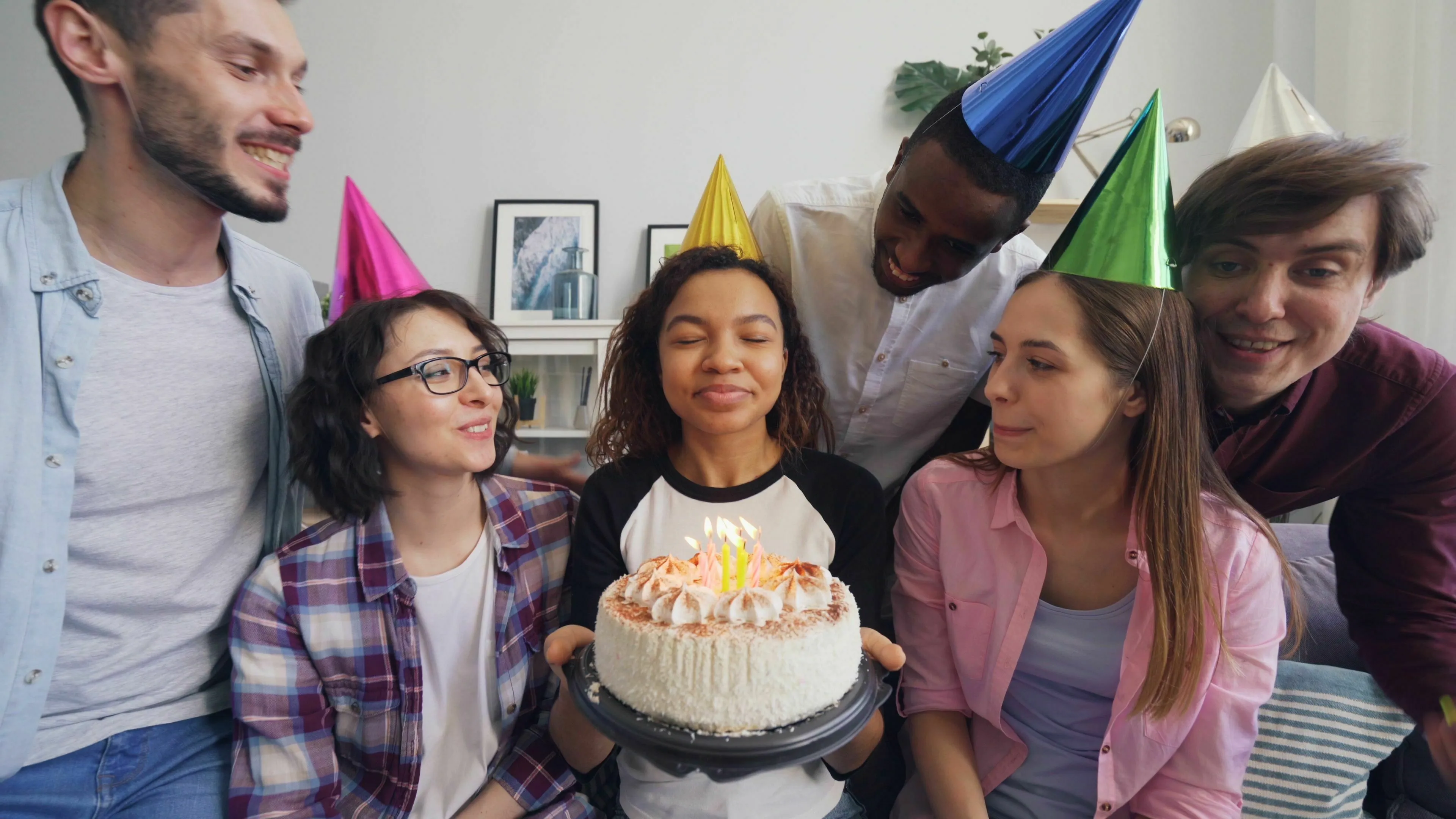 A group of diverse friends celebrates a birthday. The birthday person, a woman with closed eyes, holds a cake with candles, making a wish. Friends wear party hats, smiling, hinting at how extroverted are you.
