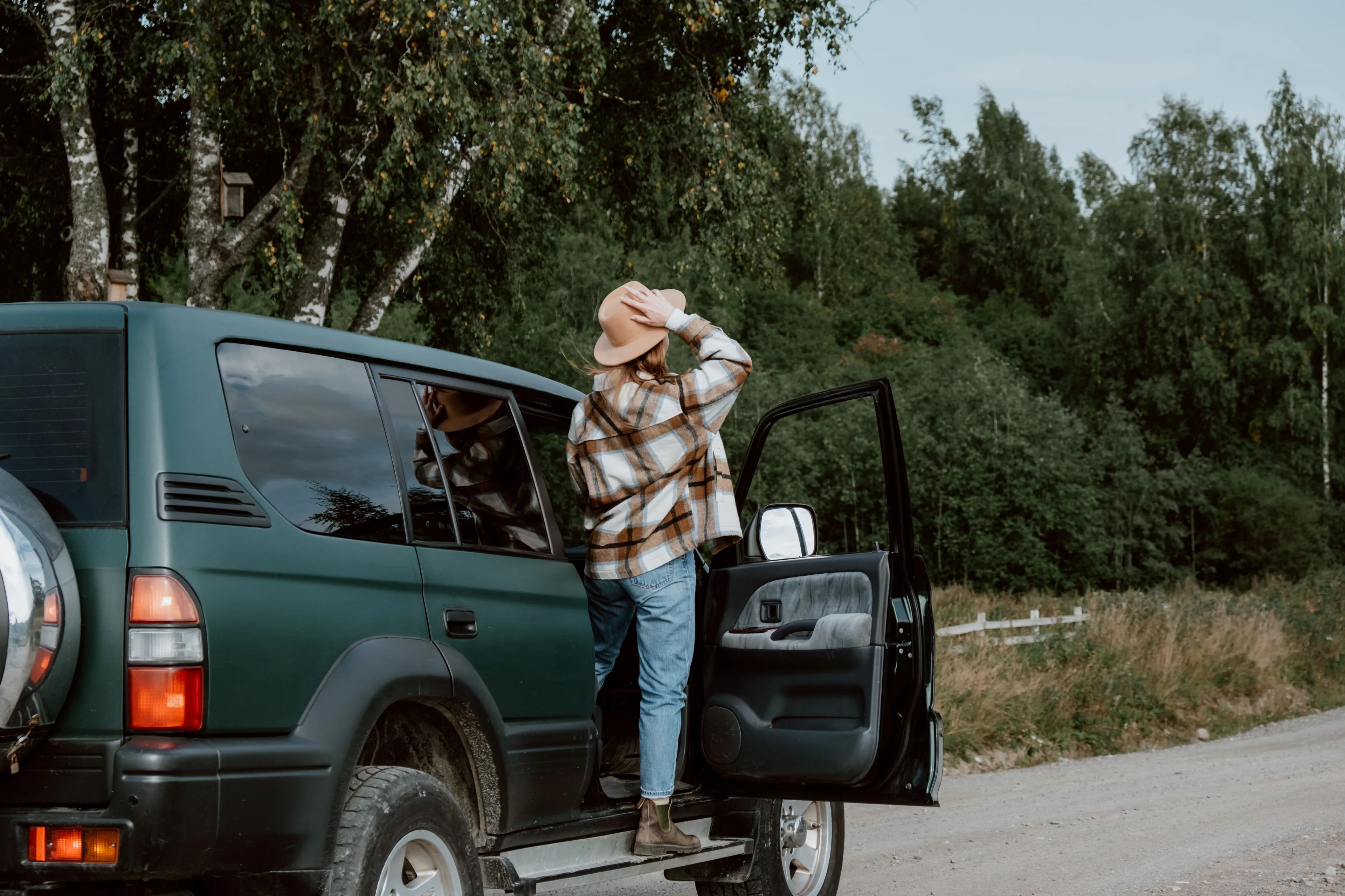 Woman in a hat and flannel shirt stepping out of a dark green SUV on a dirt road, starting a solo road trip.