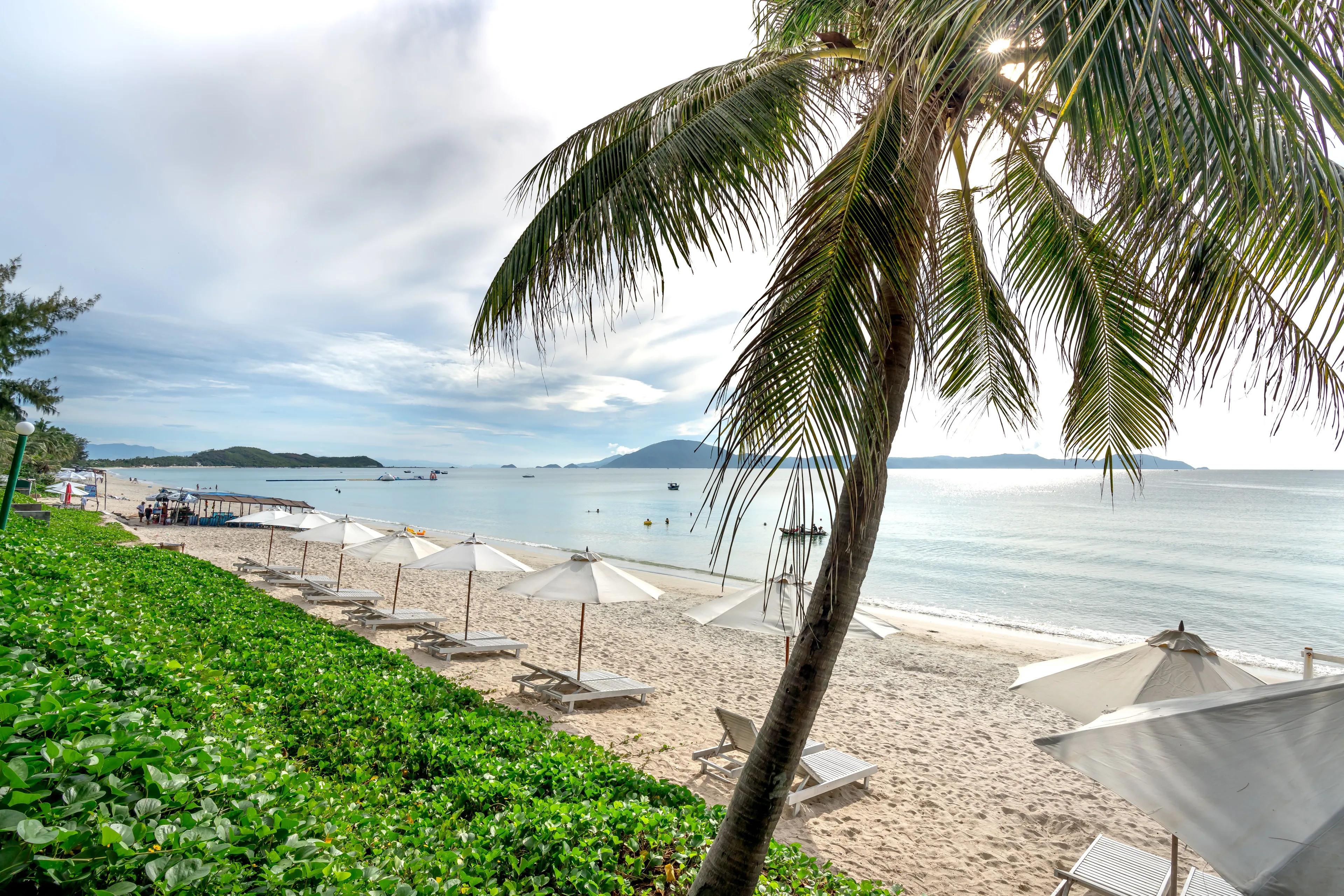 A beautiful beach resort with palm trees, white sand, and umbrellas by the calm ocean under a partly cloudy sky.