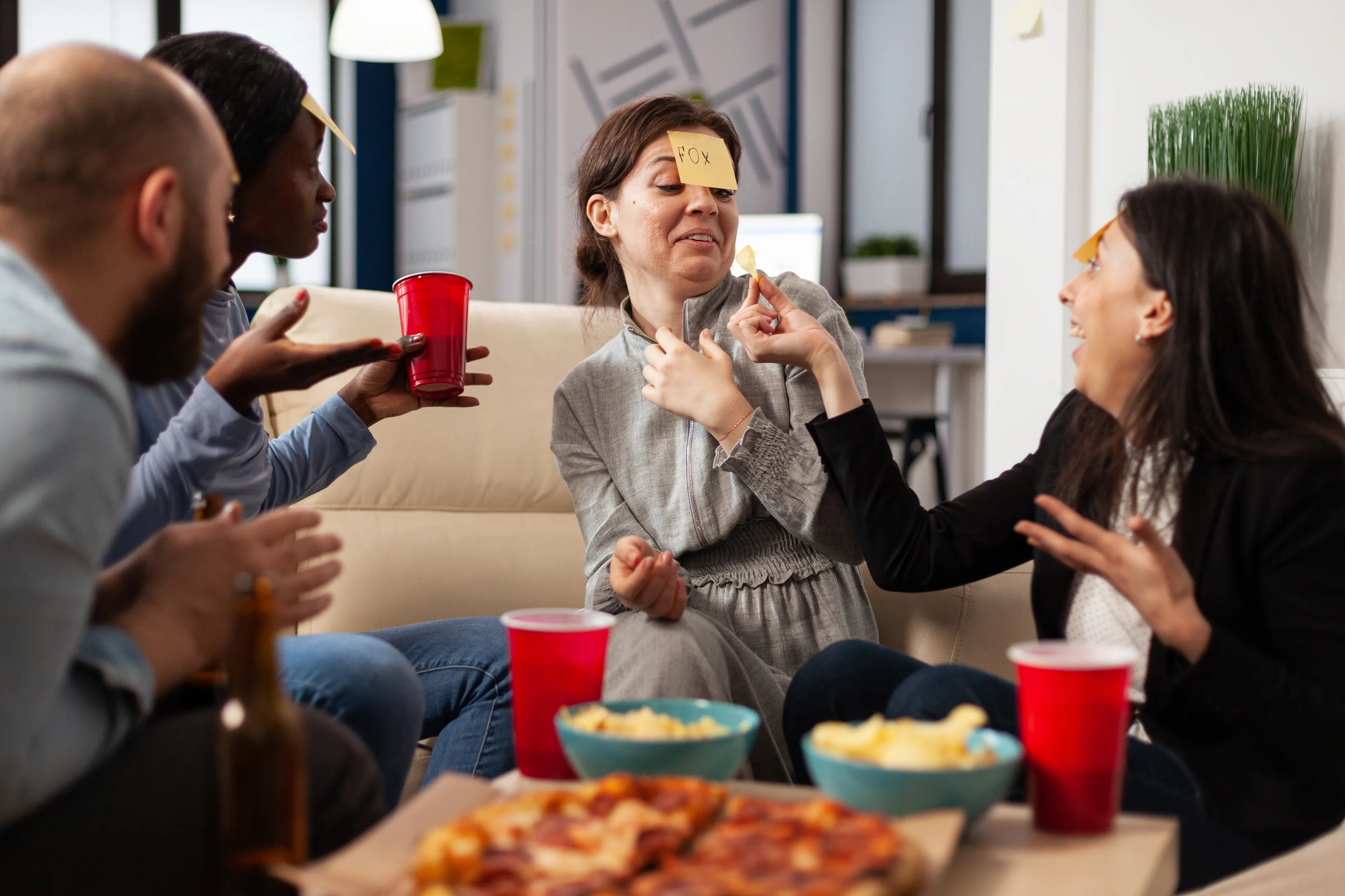 Friends enjoying a game of Charades, one woman with "FOX" on her forehead, laughing and gesturing, with pizza and snacks.
