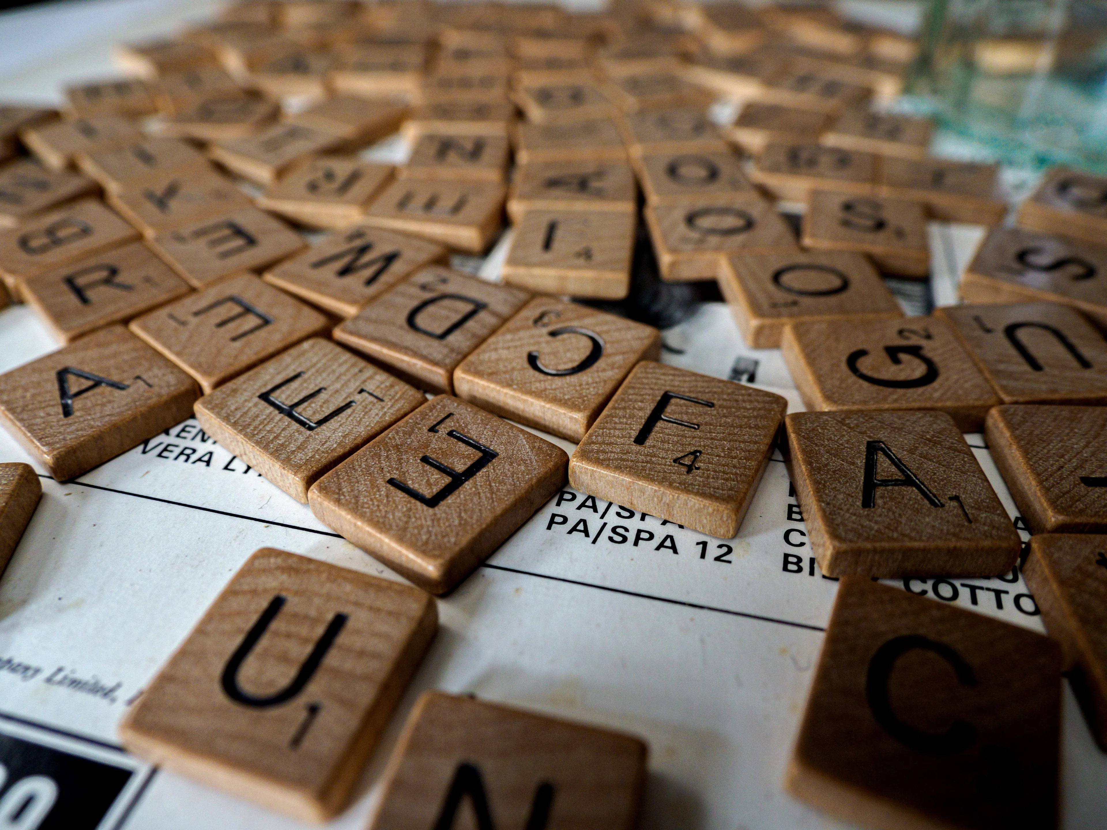 Close-up of numerous wooden Scrabble tiles scattered on a white board, featuring letters and point values.